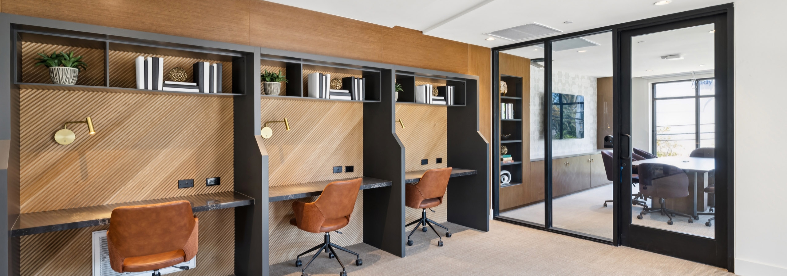 Coworking area at AMLI Aero with built-in desks, warm wood accents, leather chairs, shelving, and glass wall to private conference room.