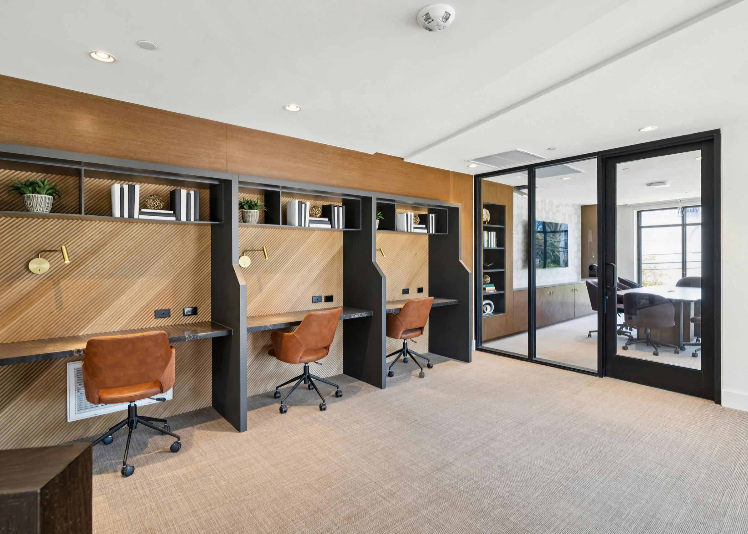 Coworking area at AMLI Aero with built-in desks, warm wood accents, leather chairs, shelving, and glass wall to private conference room.