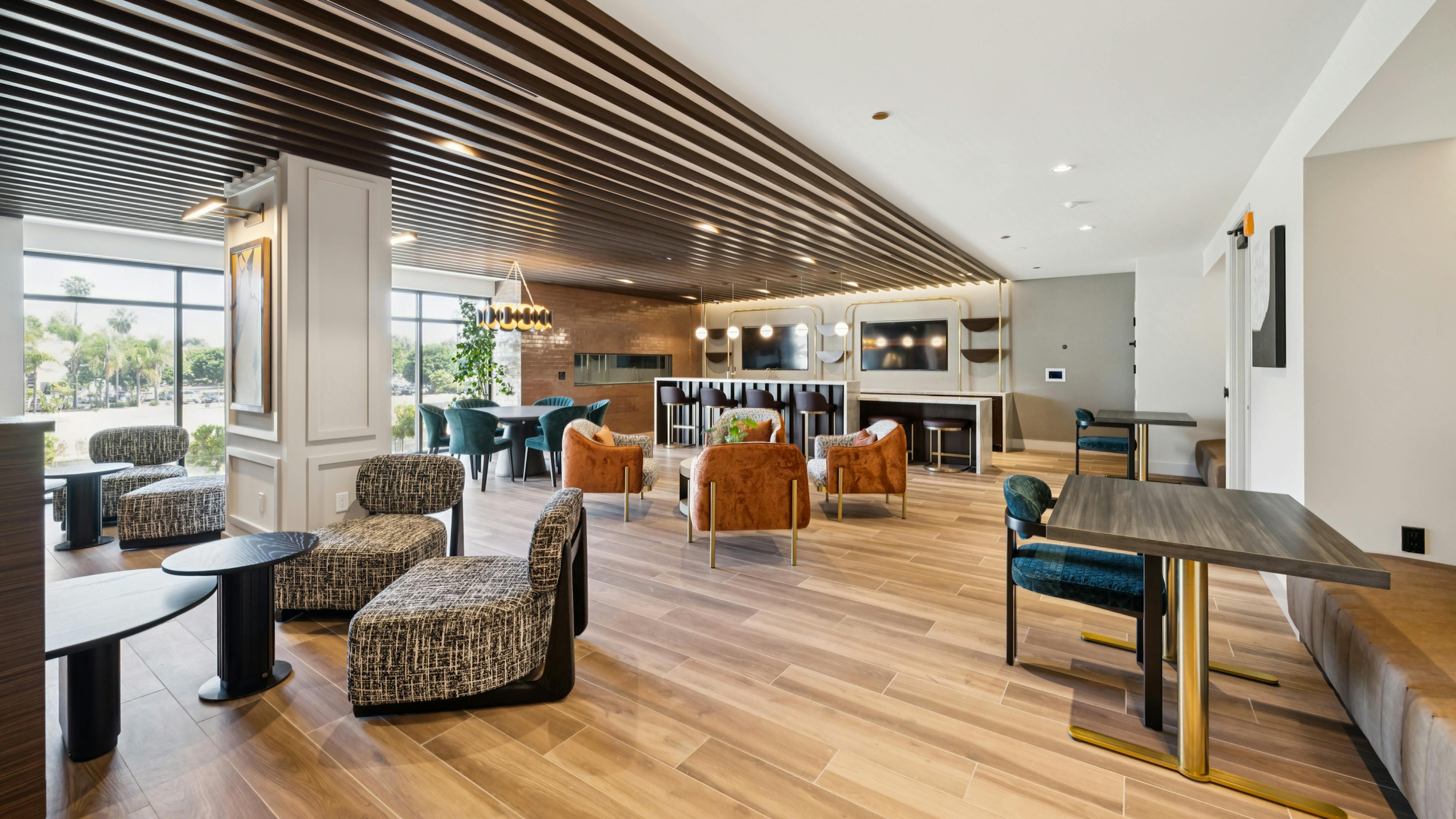 Lounge at AMLI Aero apartments with velvet seating, bar area, dining tables, wood plank floors, pendant lighting, and wall-mounted TV.