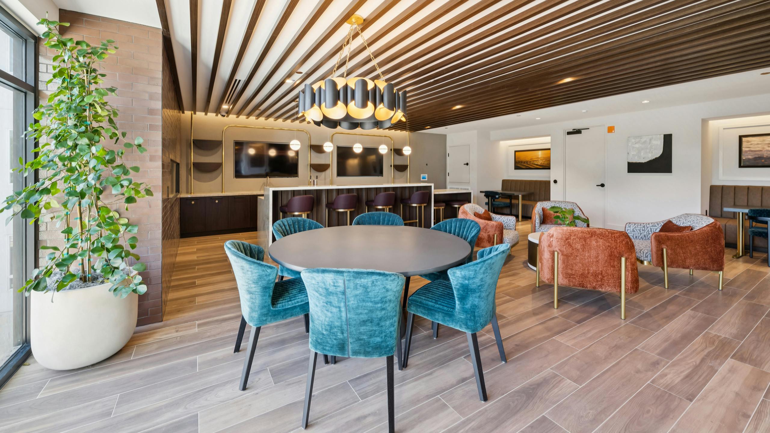 Lounge at AMLI Aero with velvet blue and orange  chairs, round dining table, wood slat ceiling, modern lighting, and two flat-screen TVs.