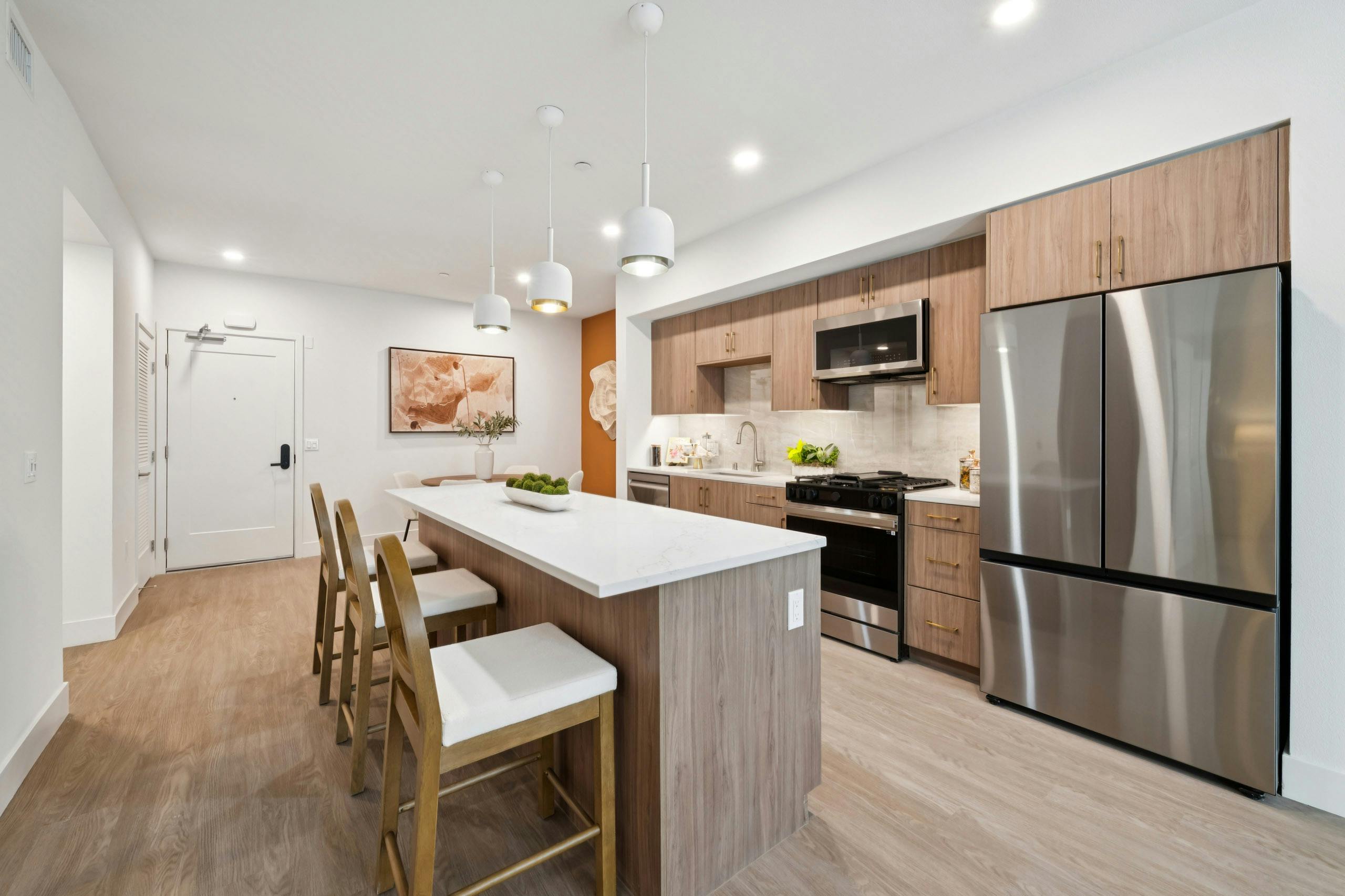 Interior view of AMLI Aero apartment kitchen with light quartz island with seating, wood-tone cabinets, gas range and peek into dining area.