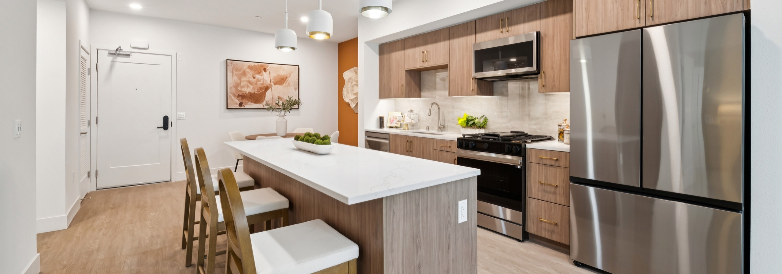 Interior view of AMLI Aero apartment kitchen with light quartz island with seating, wood-tone cabinets, gas range and peek into dining area.
