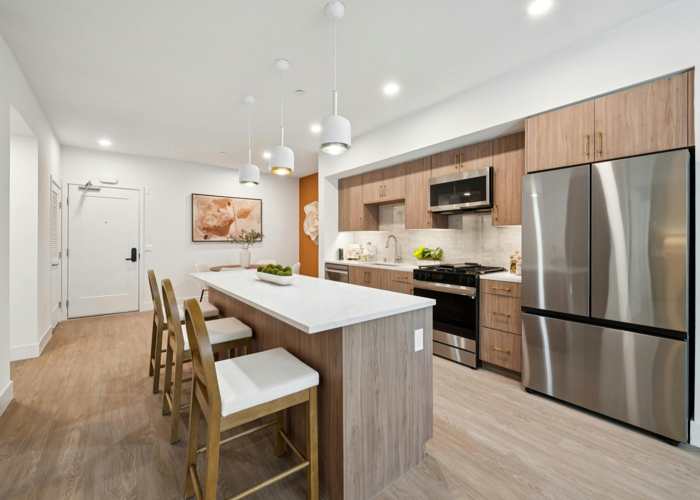 Interior view of AMLI Aero apartment kitchen with light quartz island with seating, wood-tone cabinets, gas range and peek into dining area.