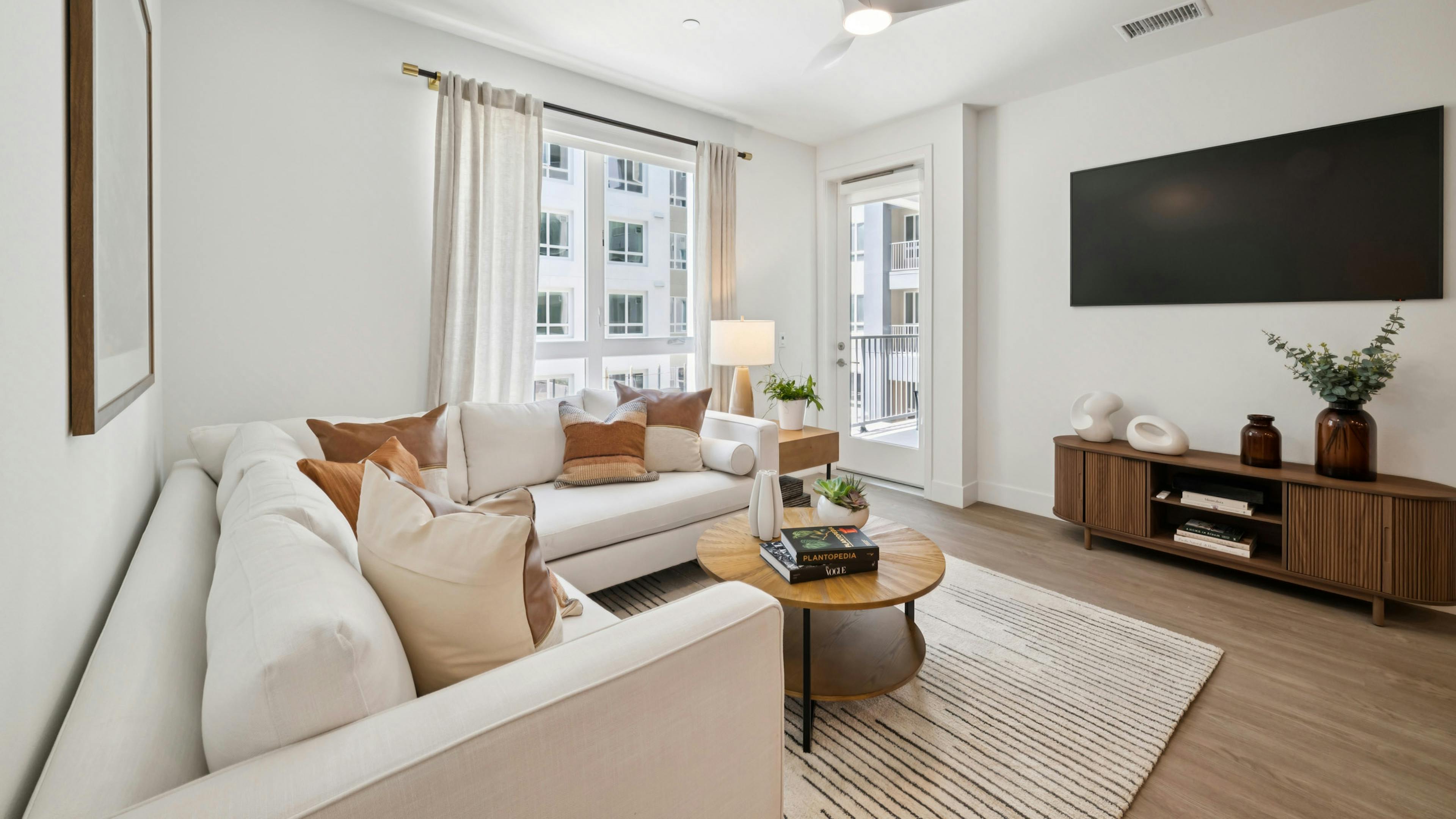 Living area  at AMLI Aero apartments featuring white sectional, accent pillows, round wood coffee table, large window, and wall mounted TV.