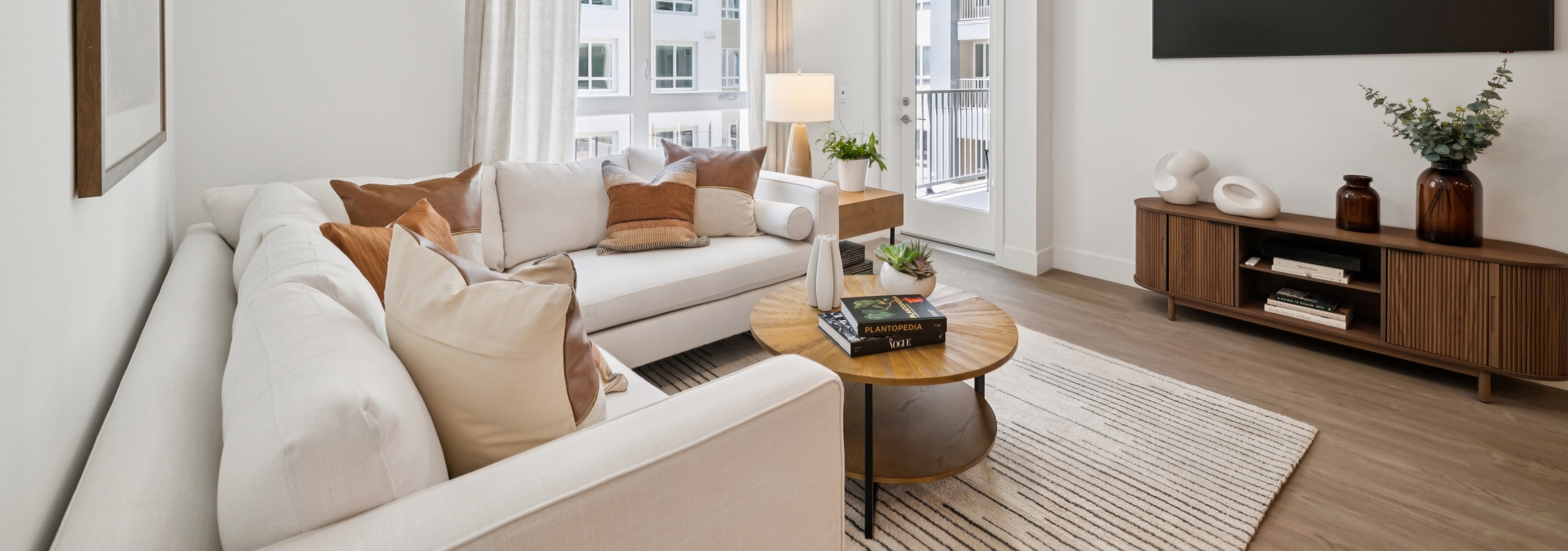 Living area  at AMLI Aero apartments featuring white sectional, accent pillows, round wood coffee table, large window, and wall mounted TV.