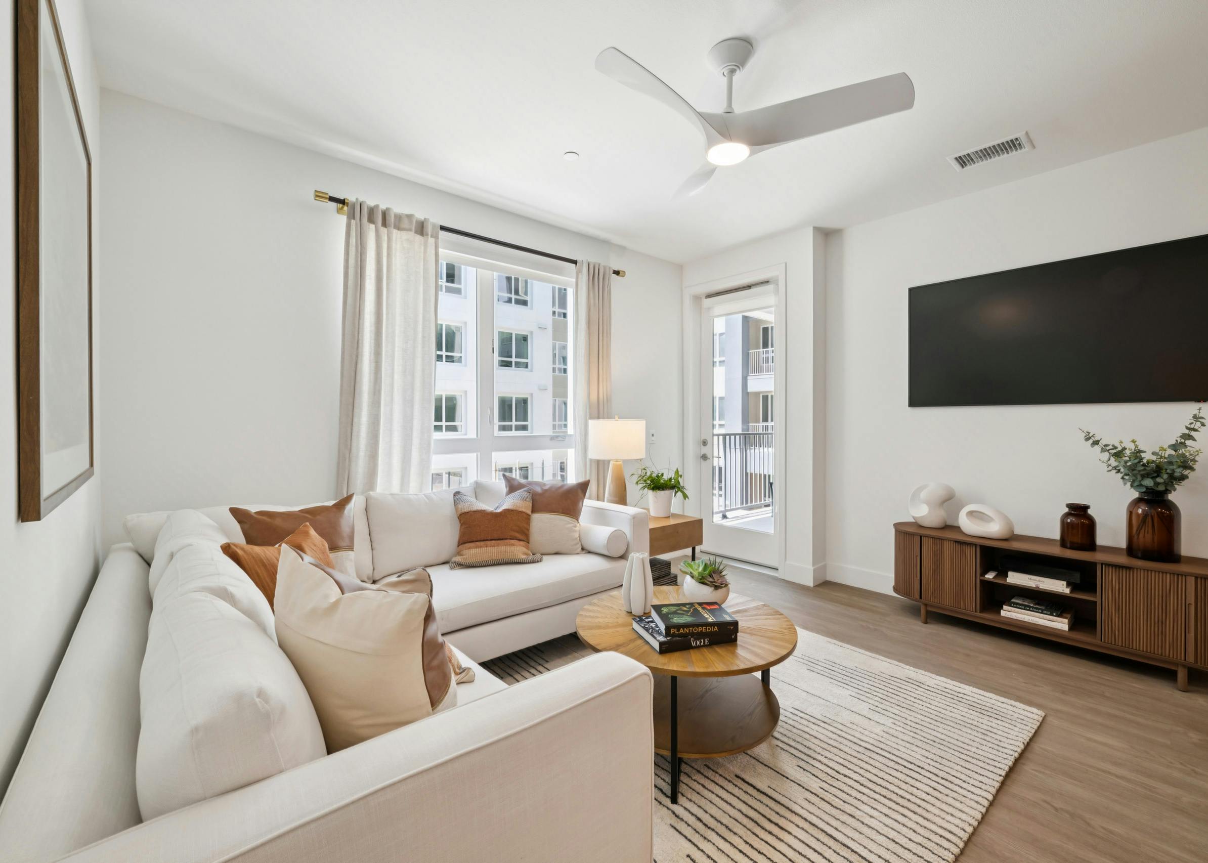 Living area at AMLI Aero apartments featuring white sectional, accent pillows, round wood coffee table, large window, and wall mounted TV.