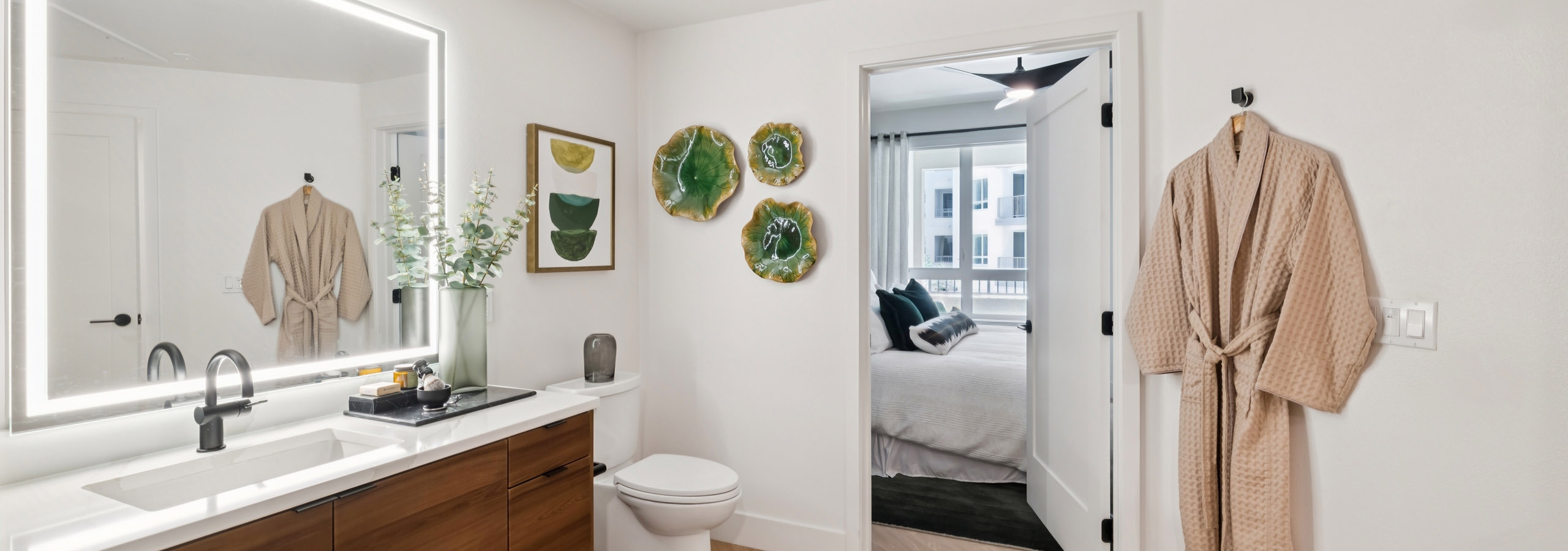 Spacious AMLI Aero apartment bathroom with warm wood vanity, oversized LED mirror, black fixtures, wall art, and open doorway to bedroom.