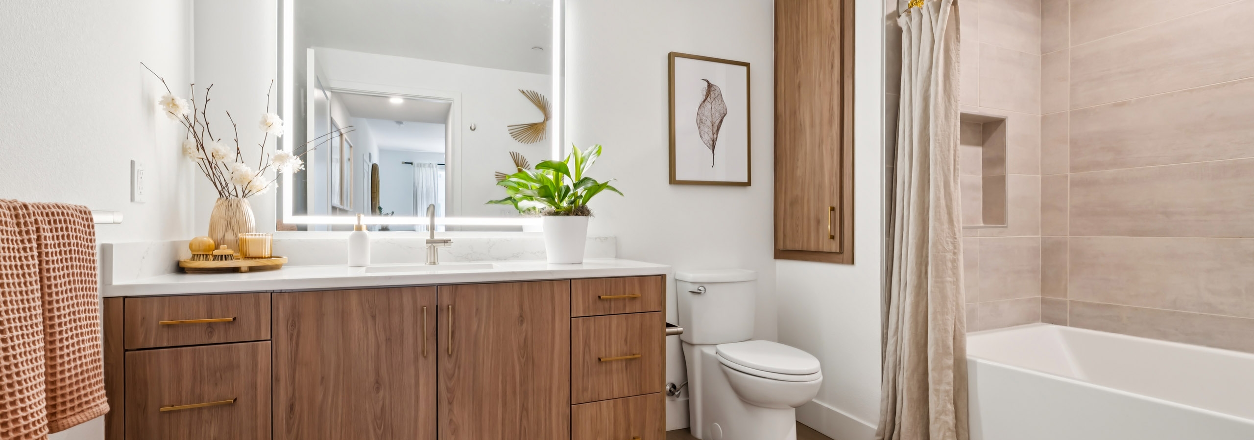 Sleek bathroom at AMLI Aero apartments with wide wood vanity, LED mirror, modern faucet, light tile shower, and brass drawer handles.