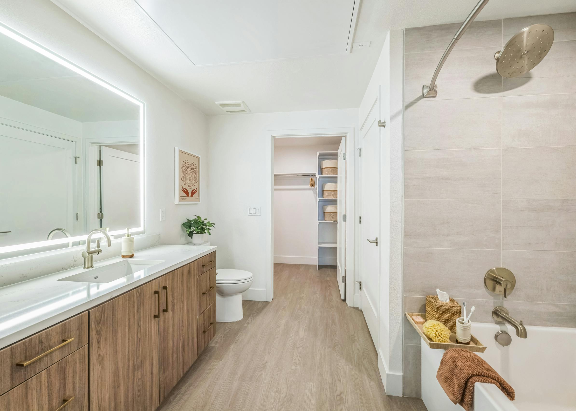 Bathroom at AMLI Aero apartments with large light wood vanity with white quartz top, LED mirror, walk-in closet, and soaking tub with tray.