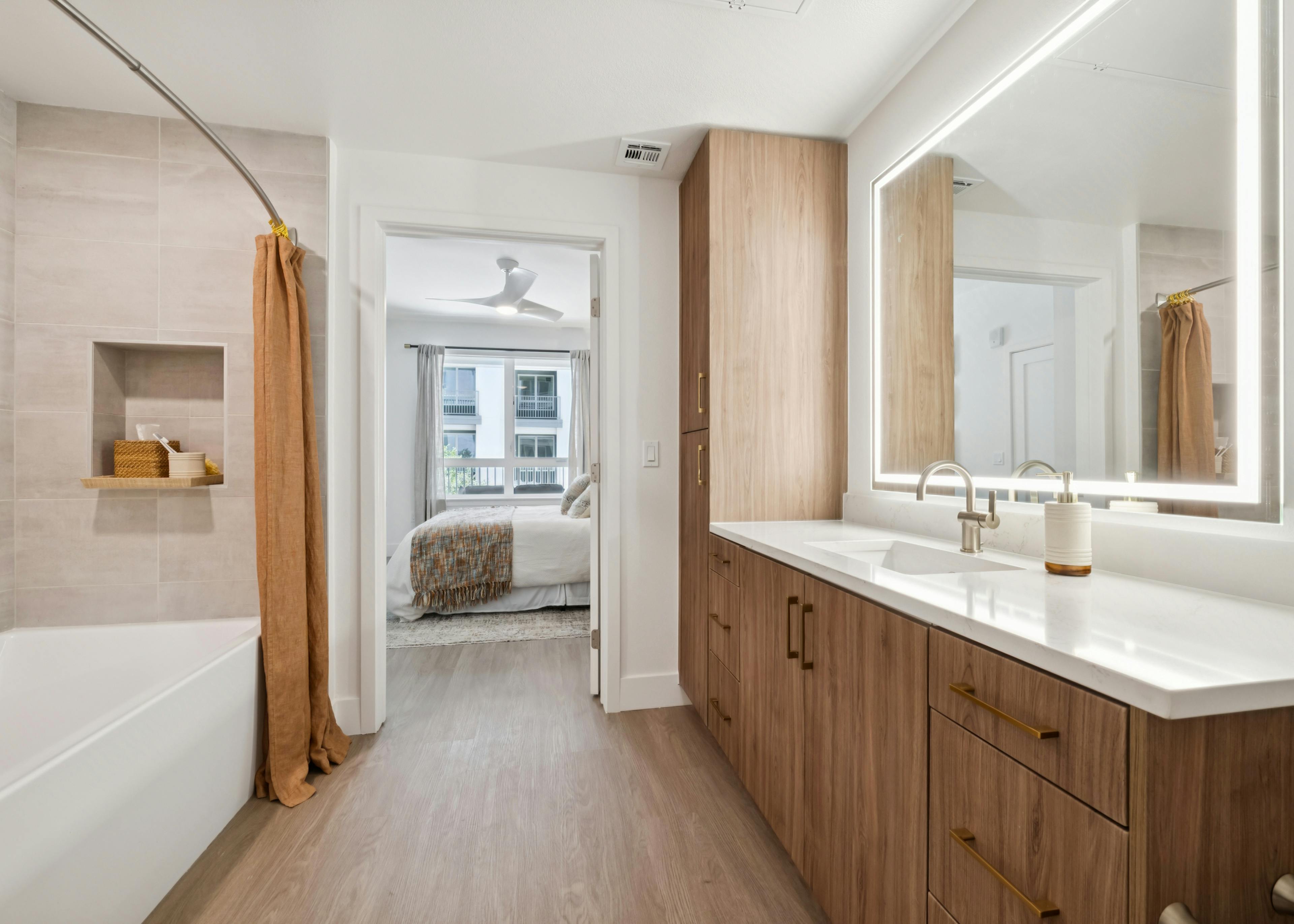 Interior view of AMLI Aero apartment bathroom with wide wood vanity, LED mirror, tile shower, and view into bedroom with ceiling fan.