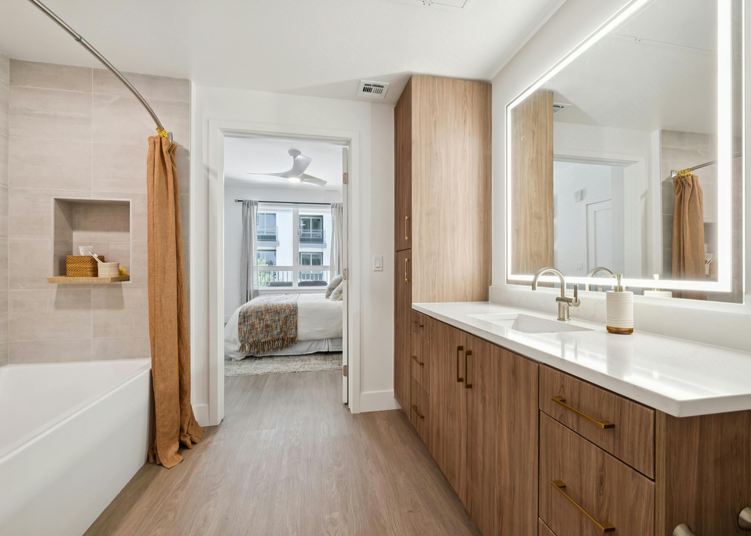 Interior view of AMLI Aero apartment bathroom with wide wood vanity, LED mirror, tile shower, and view into bedroom with ceiling fan.