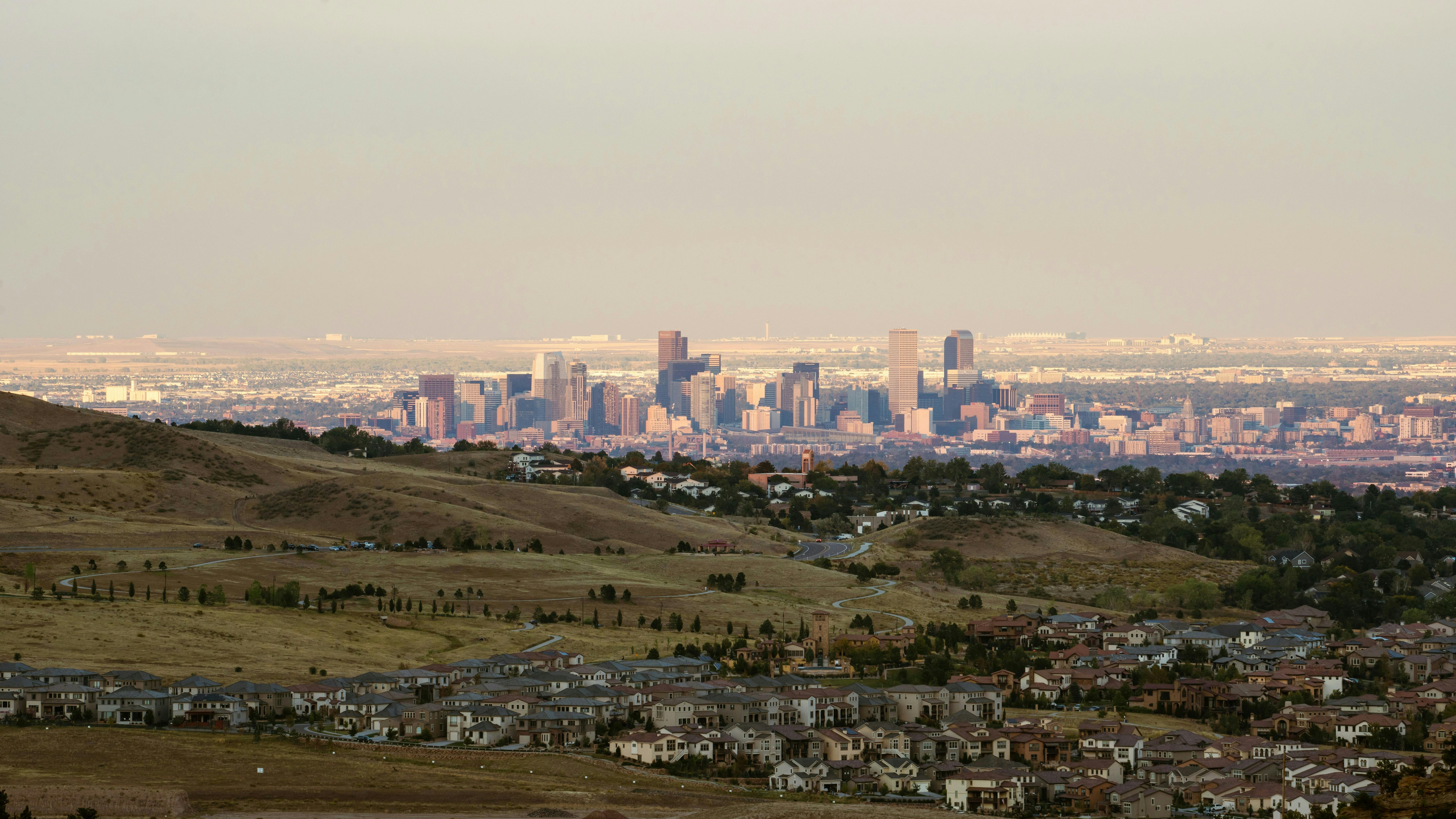 A wide view of downtown Denver, Colorado, showcasing the skyline from a distance. In the foreground, dry, rolling hills are dotted with suburban housing developments.
