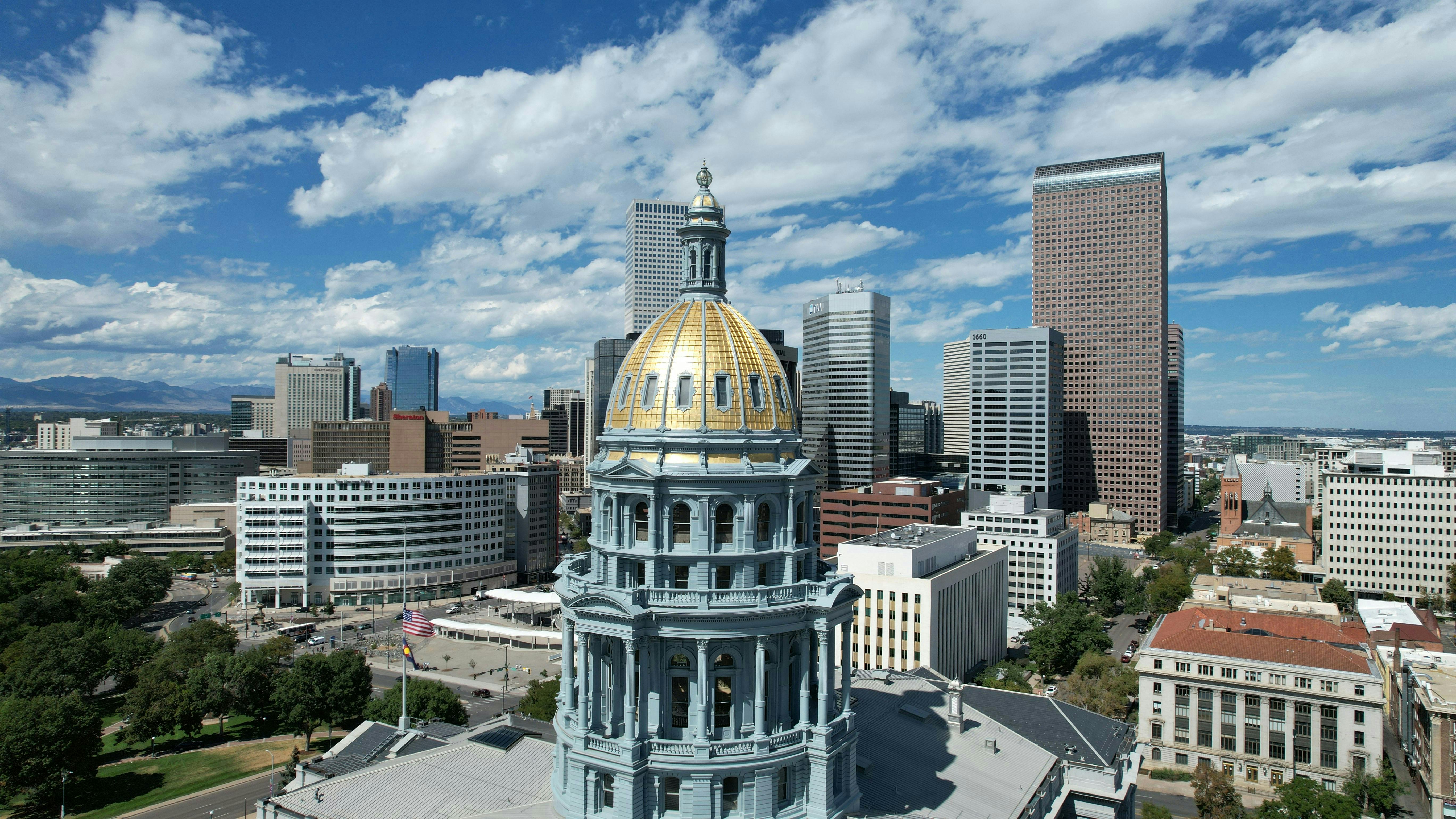 An aerial view of downtown Denver, Colorado, featuring the golden-domed Colorado State Capitol building prominently in the foreground. Modern skyscrapers rise behind it under a blue sky with fluffy white clouds, and distant mountains are visible.