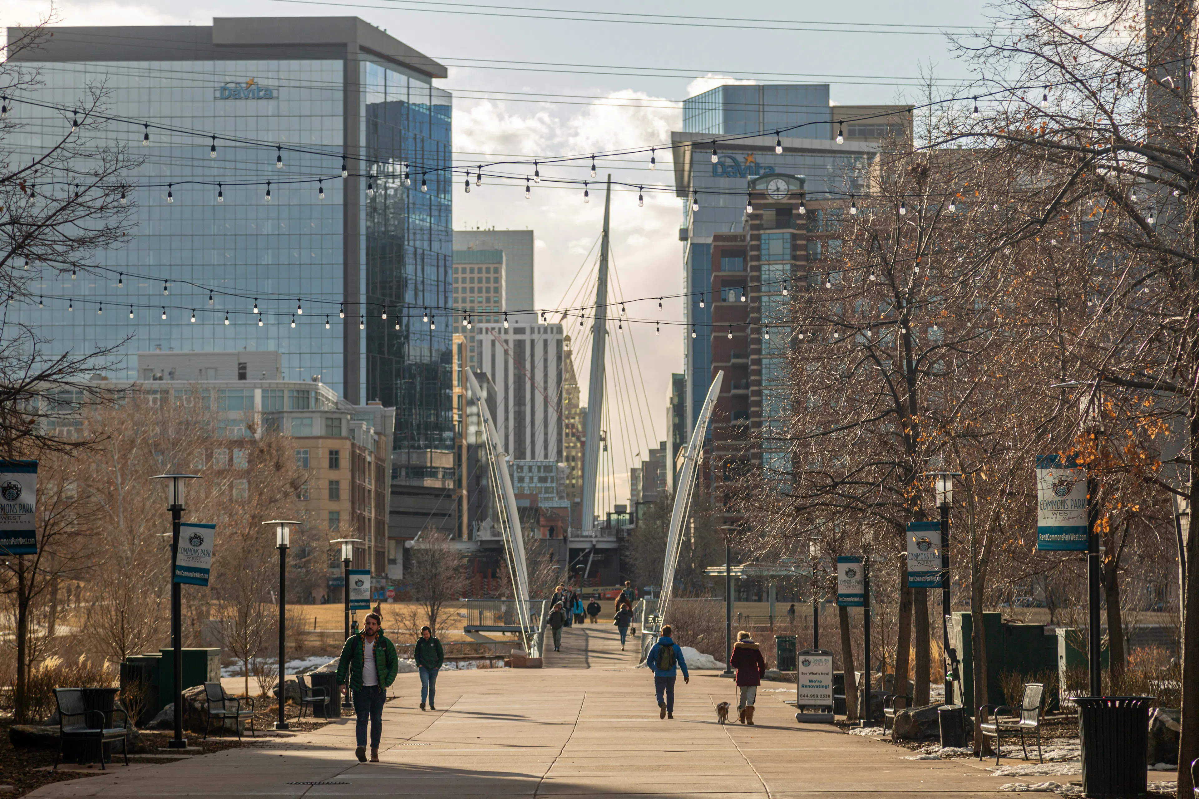 People walk along a wide, paved pathway leading towards the Millennium Bridge in Denver, Colorado. Modern glass buildings rise in the background under a bright sky, with bare trees on either side of the path strung with decorative lights.