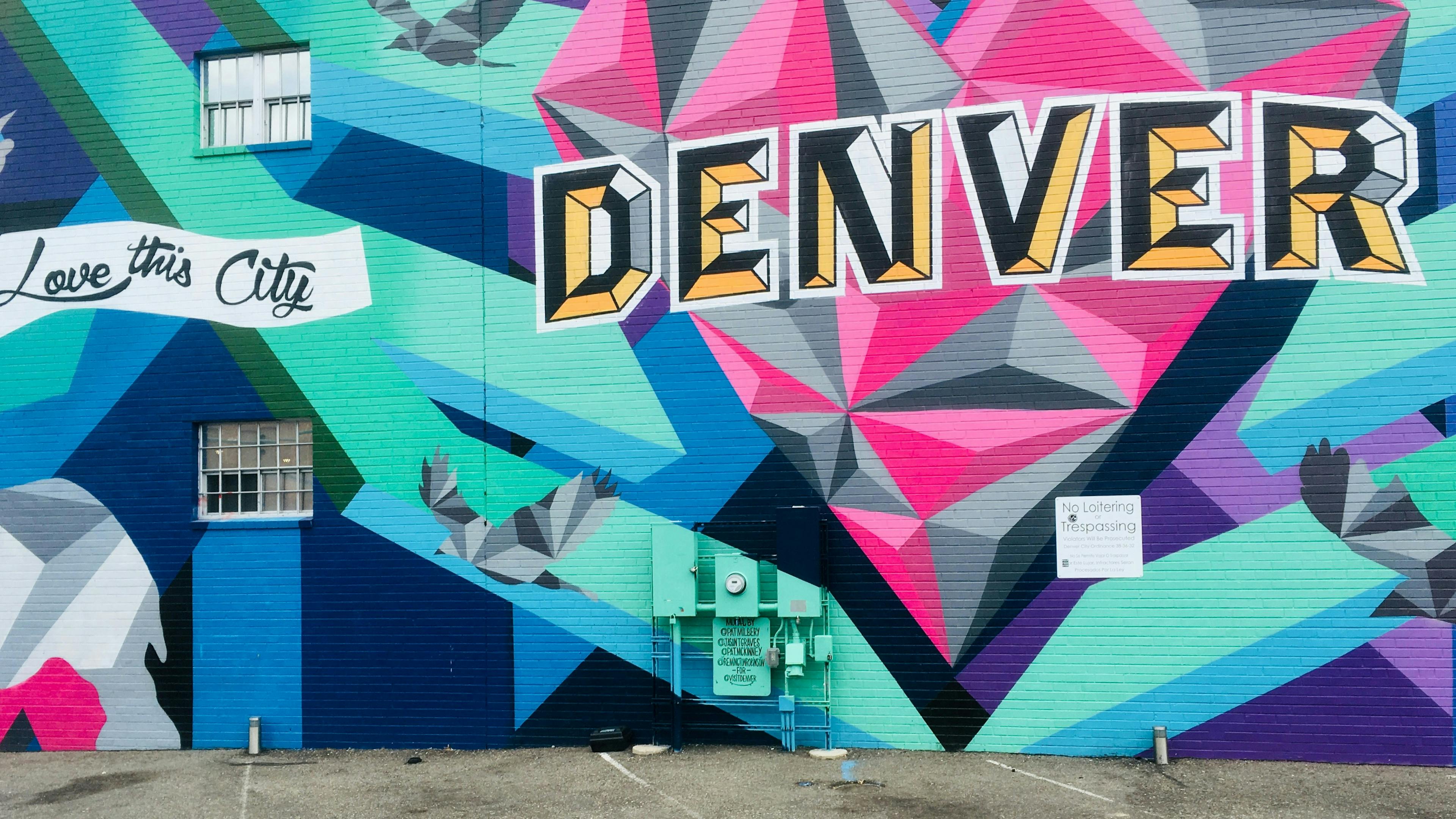 A vibrant mural on a brick wall featuring a large geometric heart design in pink, blue, and black, with the word "DENVER" in bold yellow and black letters across it. Birds in a similar geometric style fly around, and "Love this City" is written in teal.