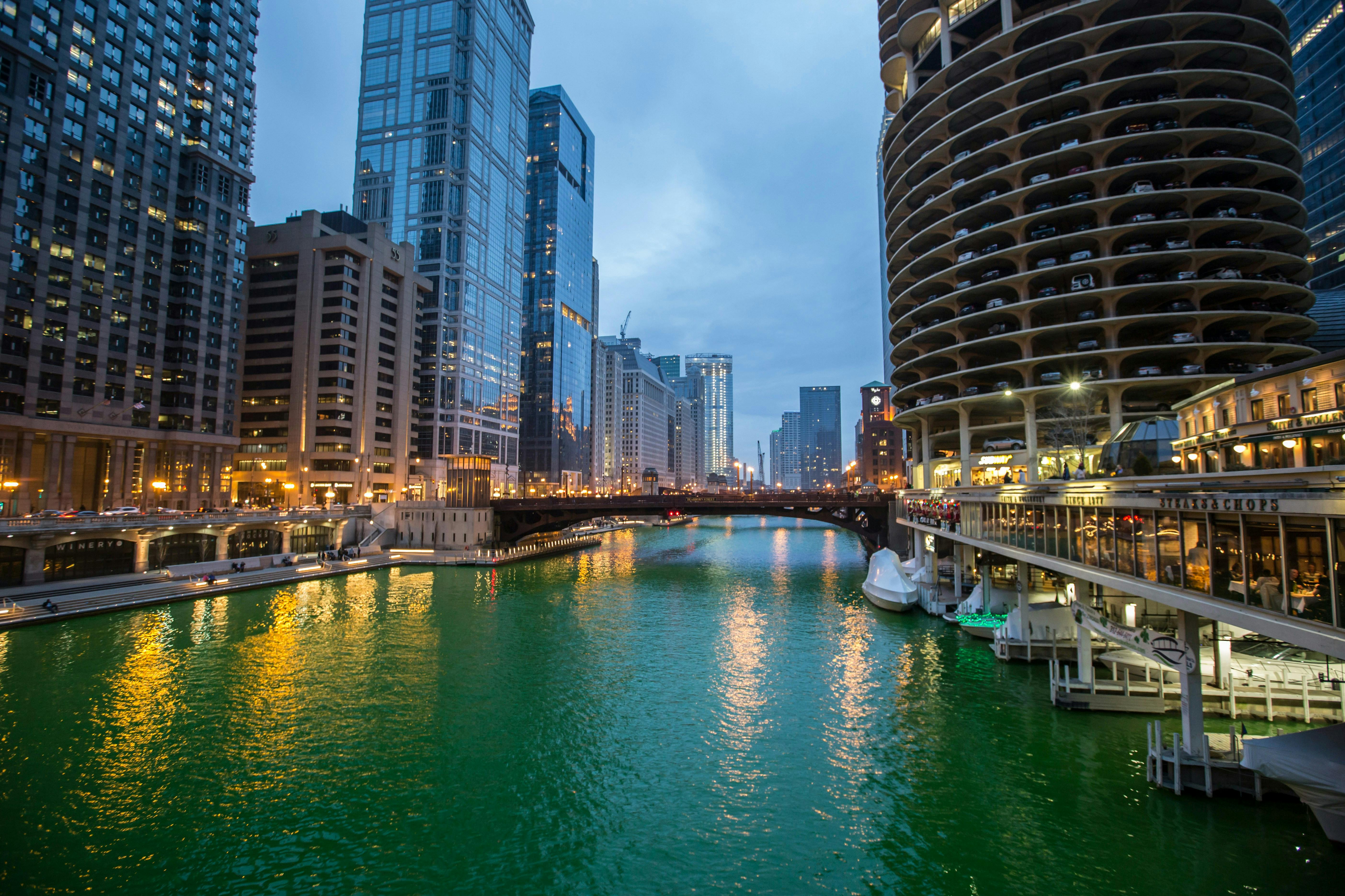 A wide shot of the Chicago River flowing between towering skyscrapers under a cloudy evening sky. The buildings are lit up, and the iconic Marina City towers are visible on the right.

