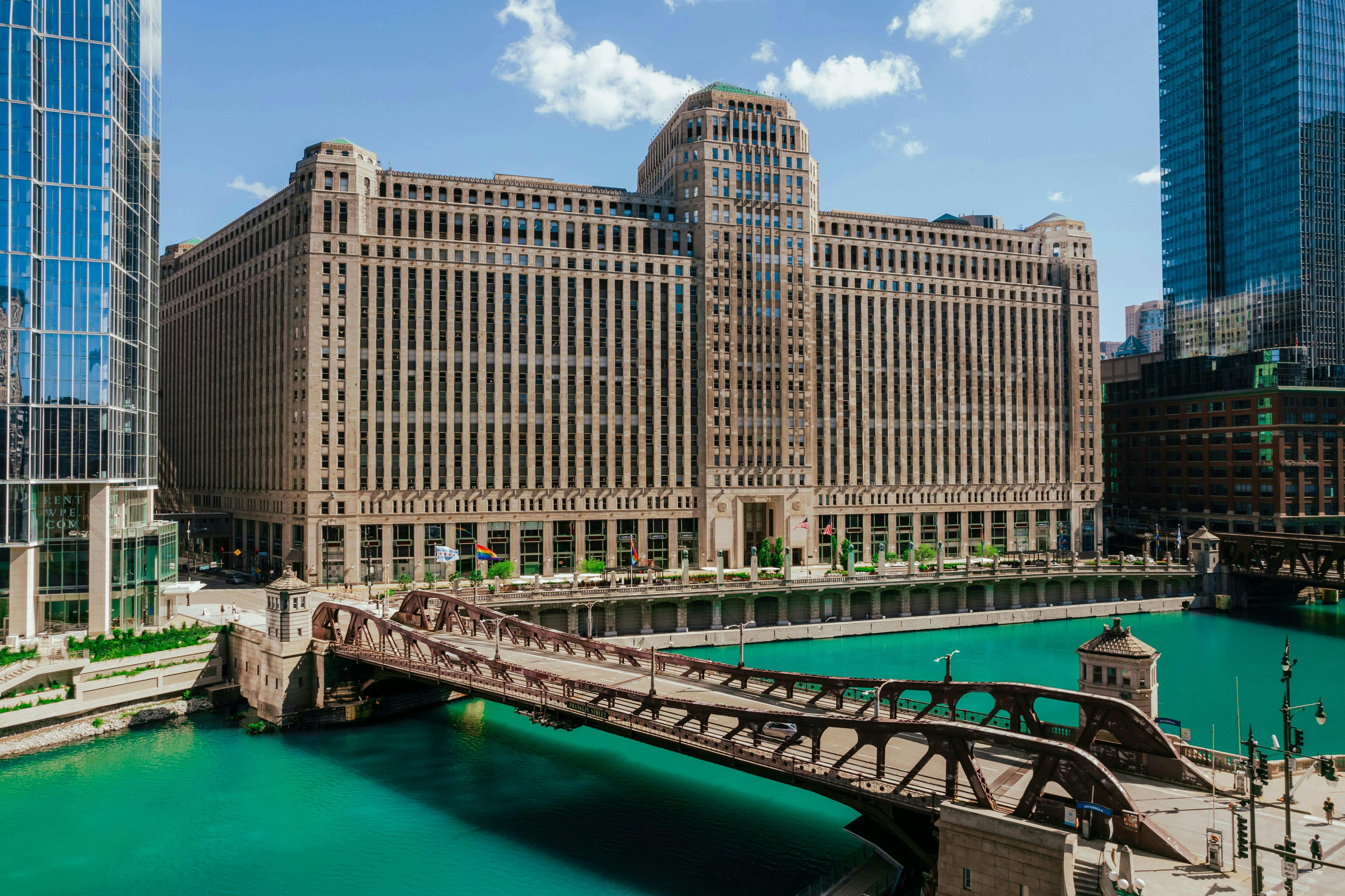 An elevated view of the Chicago River with its distinctive teal water, showing a large historic building (likely the Merchandise Mart) spanning the river, connected by a drawbridge. Modern glass skyscrapers flank the river under a partly cloudy sky.