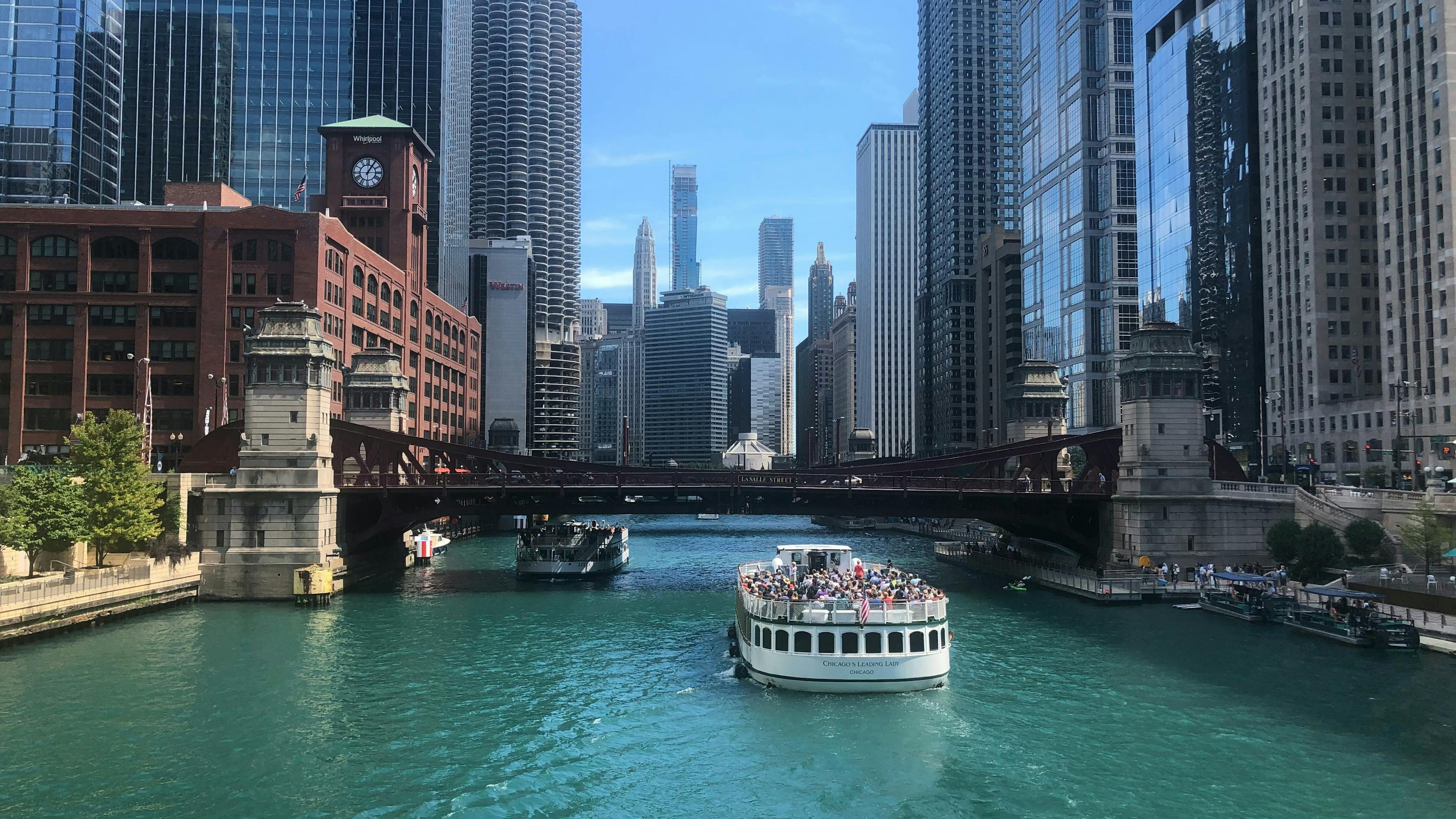 A bustling scene on the Chicago River, with a large tourist boat filled with people cruising on the teal-colored water. High-rise buildings and bridges span the river under a bright blue sky.