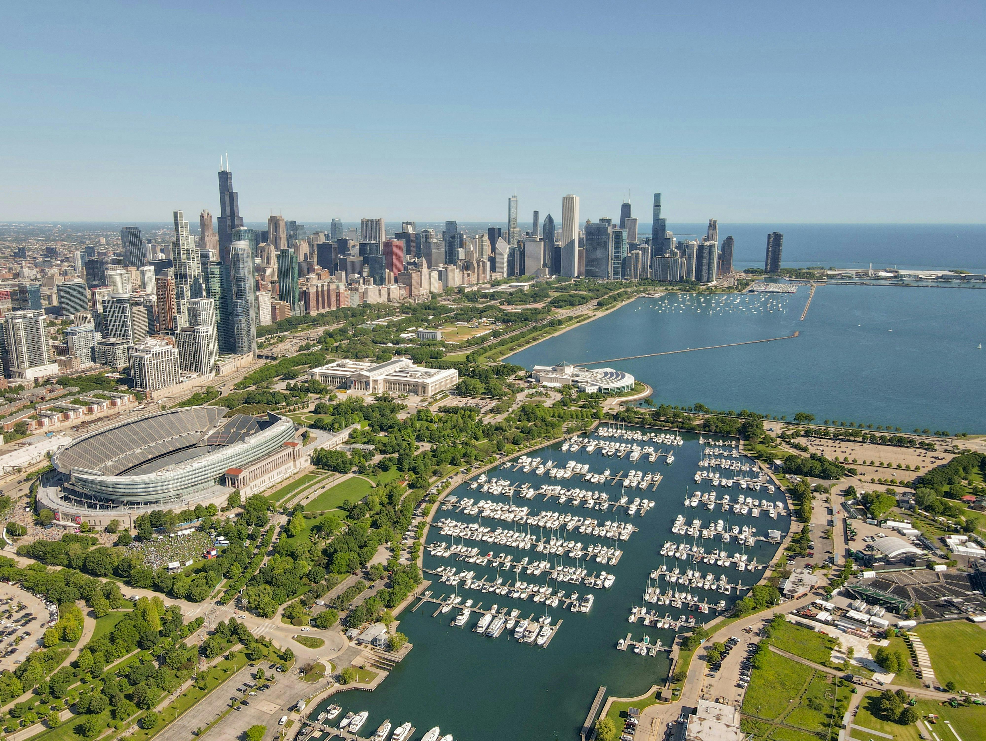 An aerial view of the Chicago skyline, showing numerous skyscrapers, Soldier Field stadium, and a large marina filled with boats on Lake Michigan under a clear blue sky.