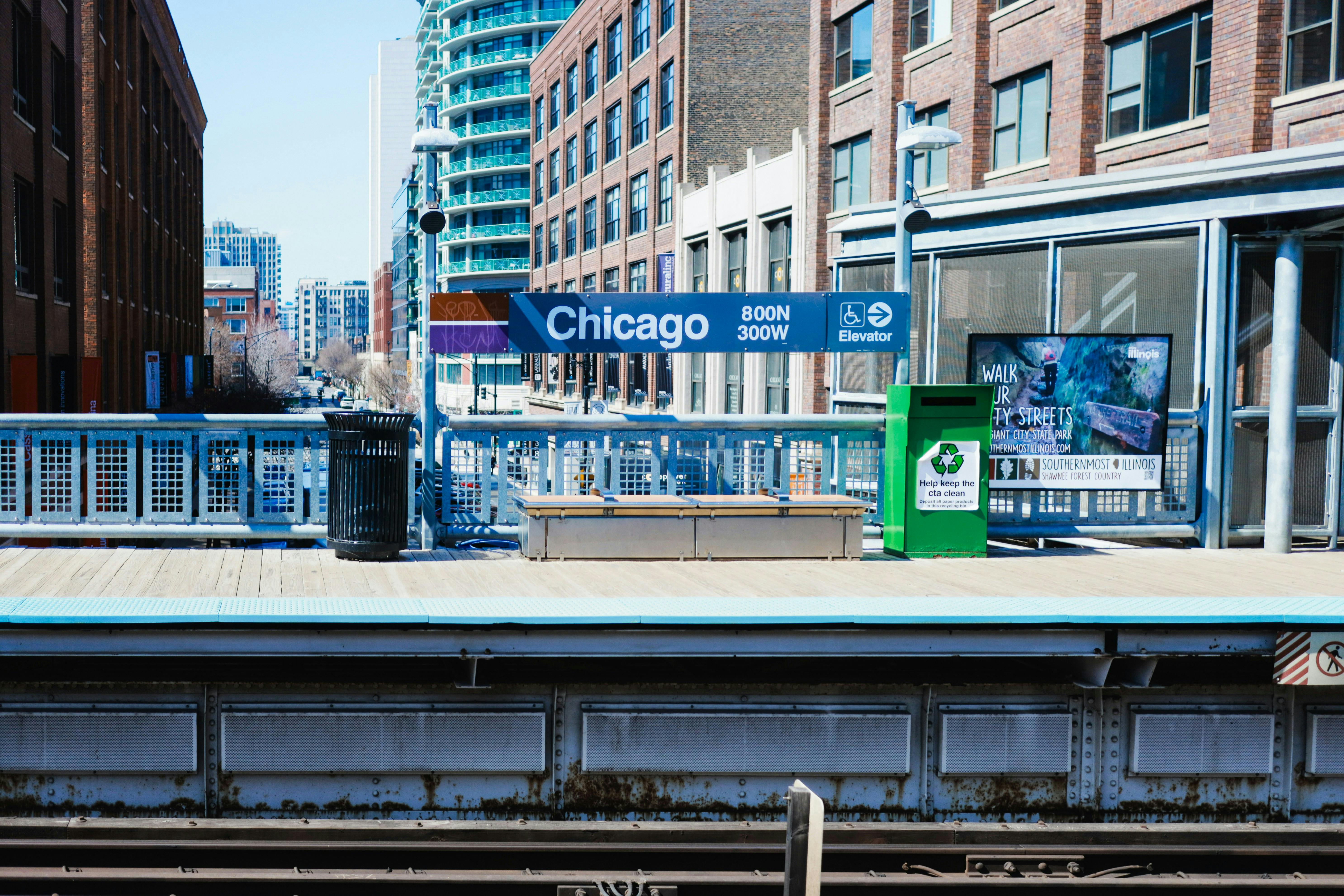 A view of a Chicago train platform with a "Chicago" sign prominently displayed. Buildings and elevated train tracks are visible in the background on a sunny day.

