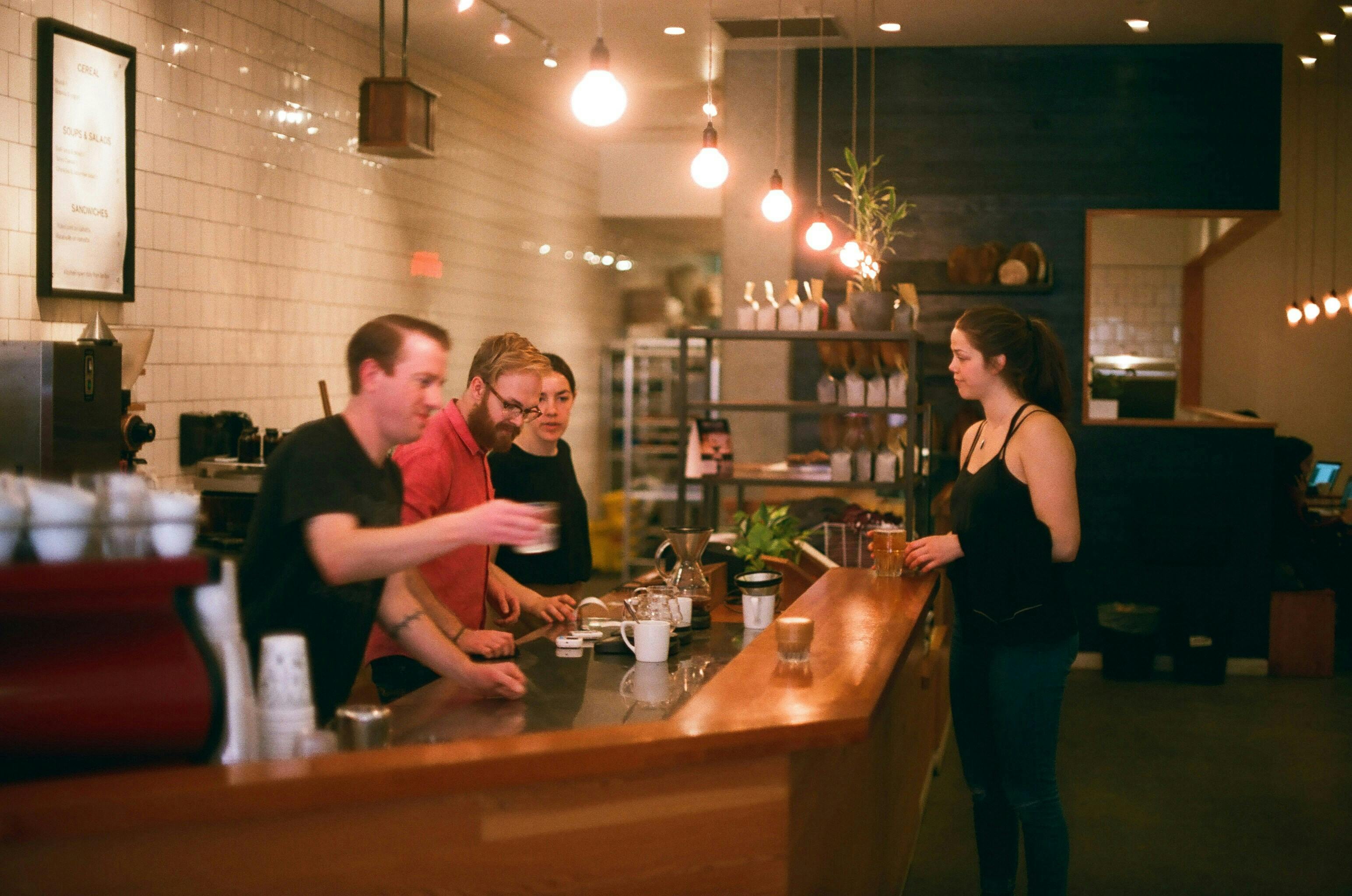 Three baristas are at a counter with a customer. The customer, a woman in a black tank top, is making an order while a man in a red shirt gestures to her.