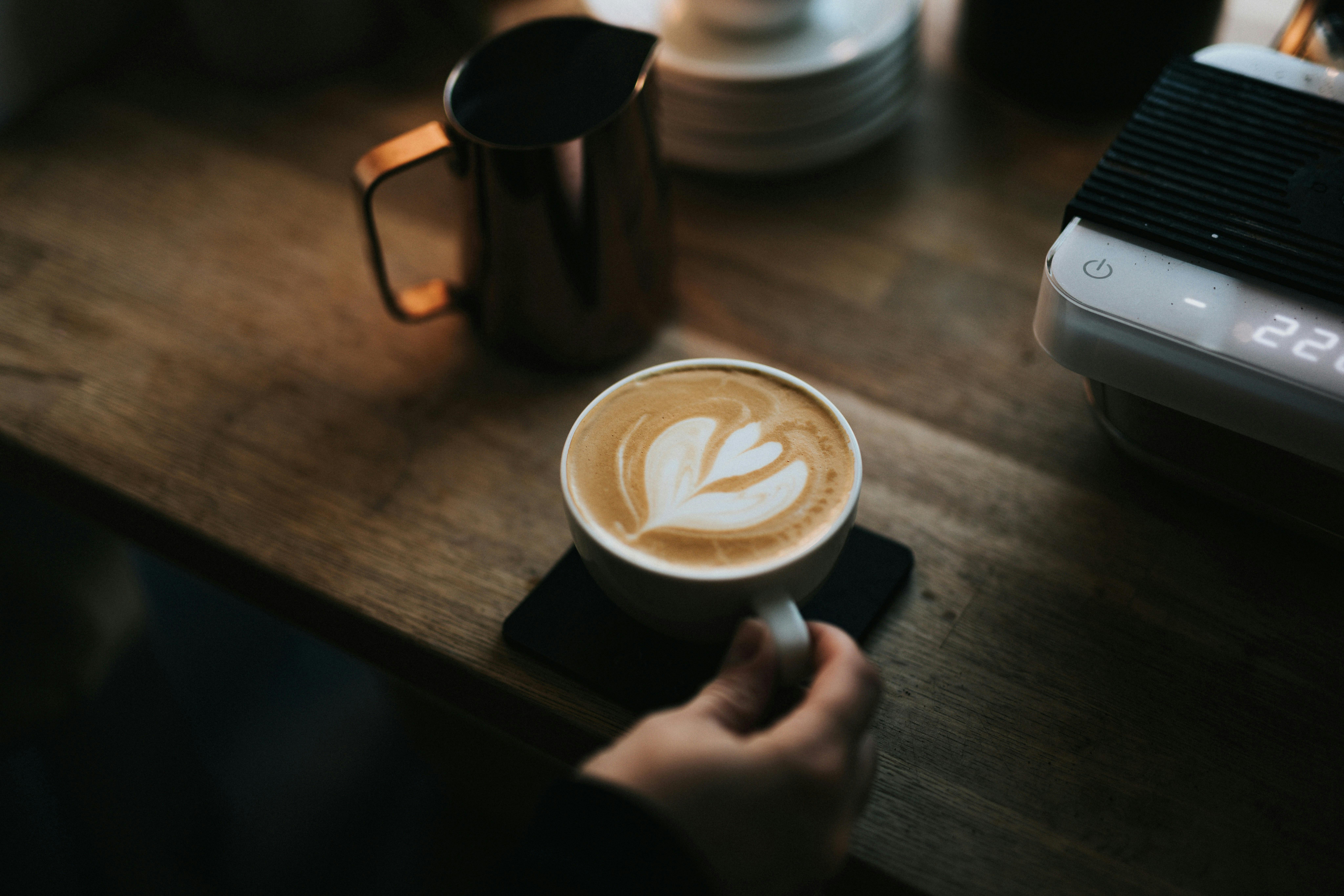 A close-up shot of a latte with heart-shaped foam art in a white mug, held by a hand. A shiny silver milk pitcher and a coffee scale are visible on a wooden counter in the background.