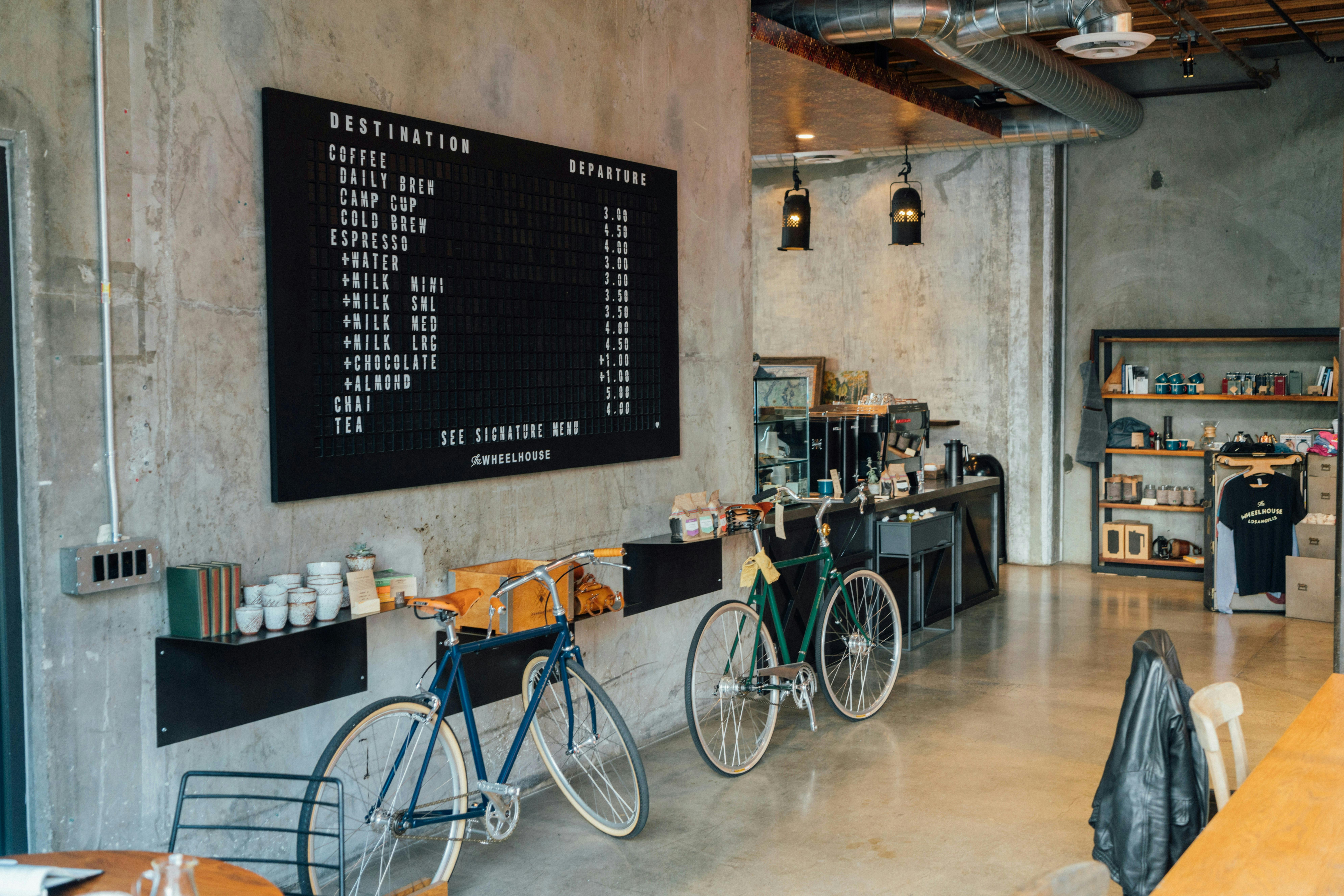The interior of a modern, industrial-style cafe with concrete walls and floors. A large black menu board on the left wall lists "DESTINATION" coffees and other drinks with their prices under "DEPARTURE." Below the menu are shelves holding mugs and books, and two bicycles, one blue and one green, are leaning against the wall. The cafe has hanging industrial lights, a service counter in the back, and shelves with merchandise.