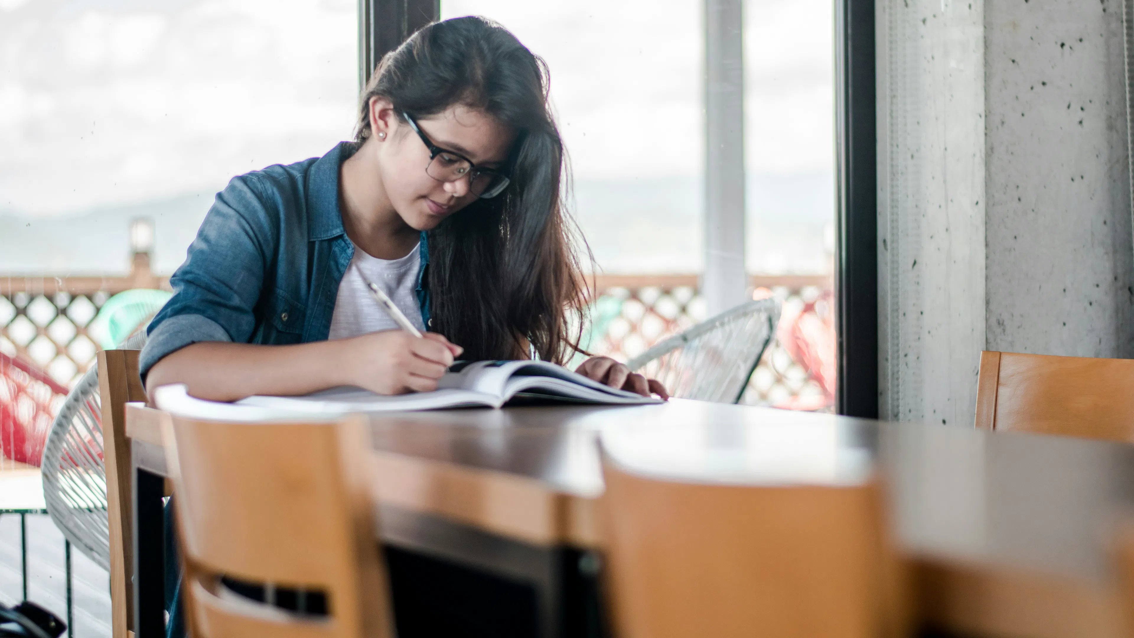 A young woman with dark hair, wearing glasses and a denim shirt over a white t-shirt, sits at a wooden table in front of a window. She is focused on writing in an open notebook with a pen.