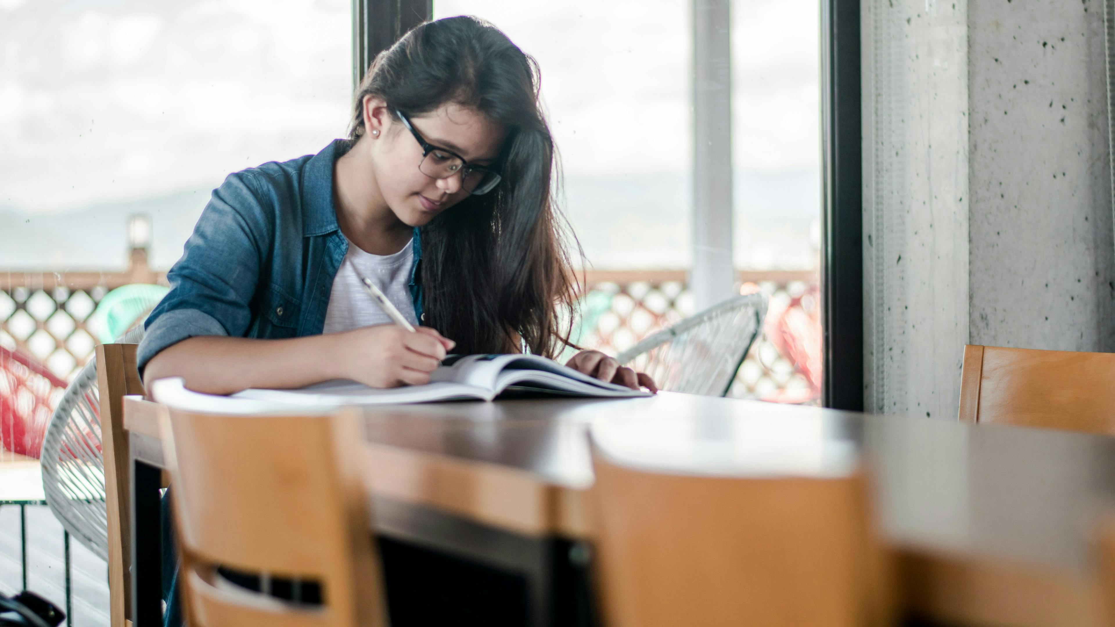 A young woman with dark hair, wearing glasses and a denim shirt over a white t-shirt, sits at a wooden table in front of a window. She is focused on writing in an open notebook with a pen.