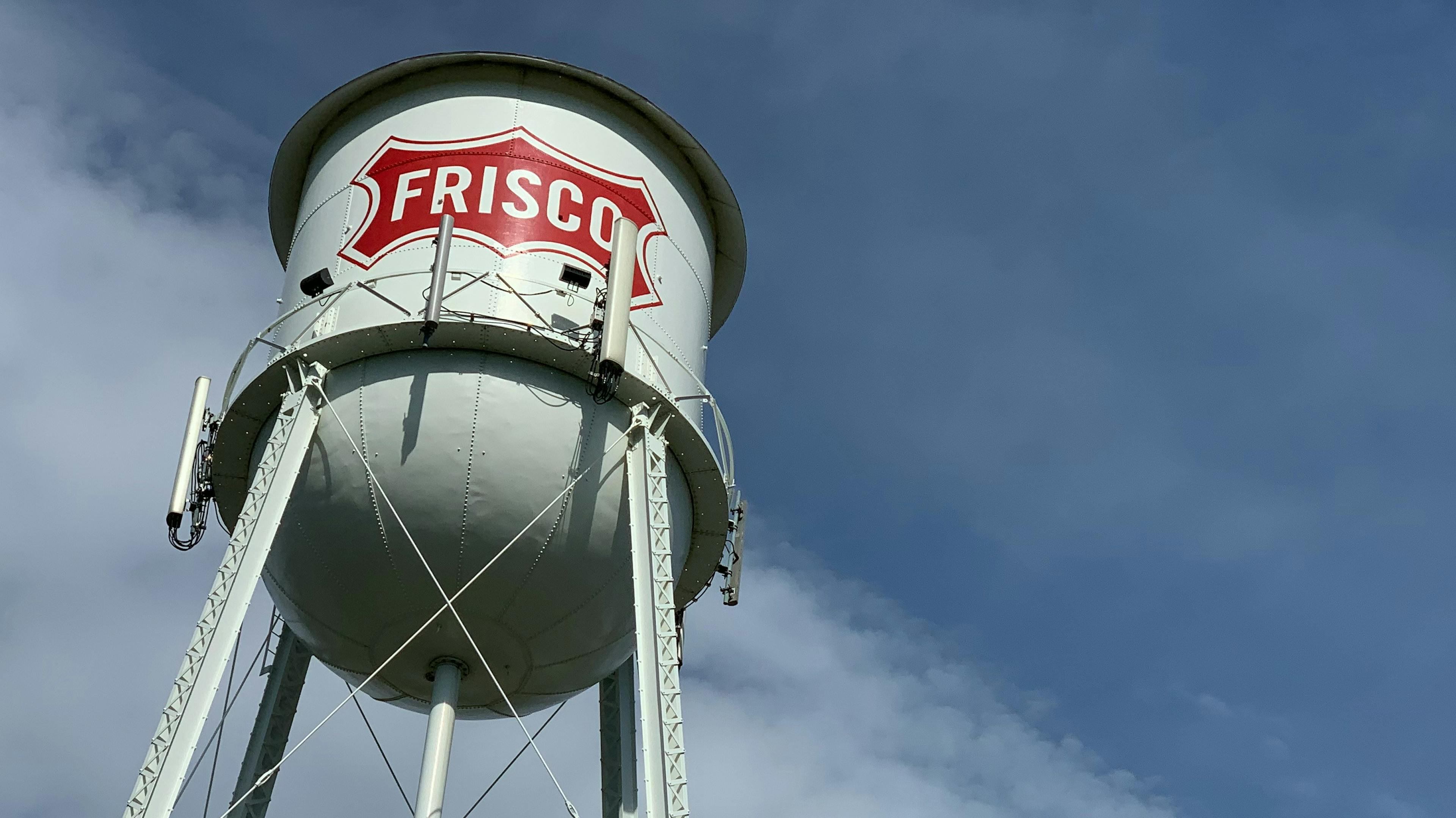 A large white water tower with the word Frisco in a bright red logo on it, set against a blue sky dotted with wispy white clouds