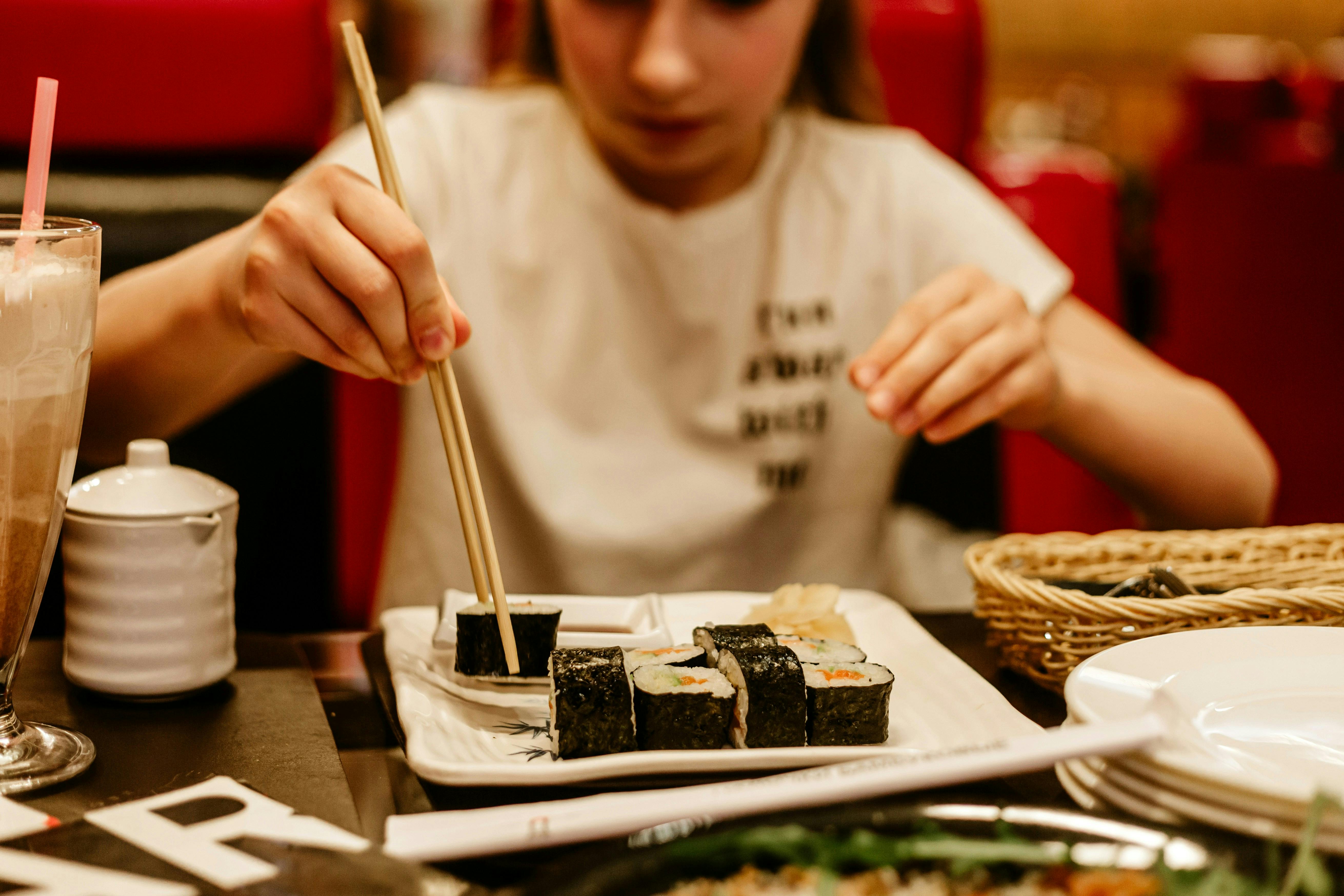 A young girl is sitting at a restaurant table and using chopsticks to pick up a piece of sushi from a rectangular plate. A drink with a pink straw and other dishes are visible on the table. 