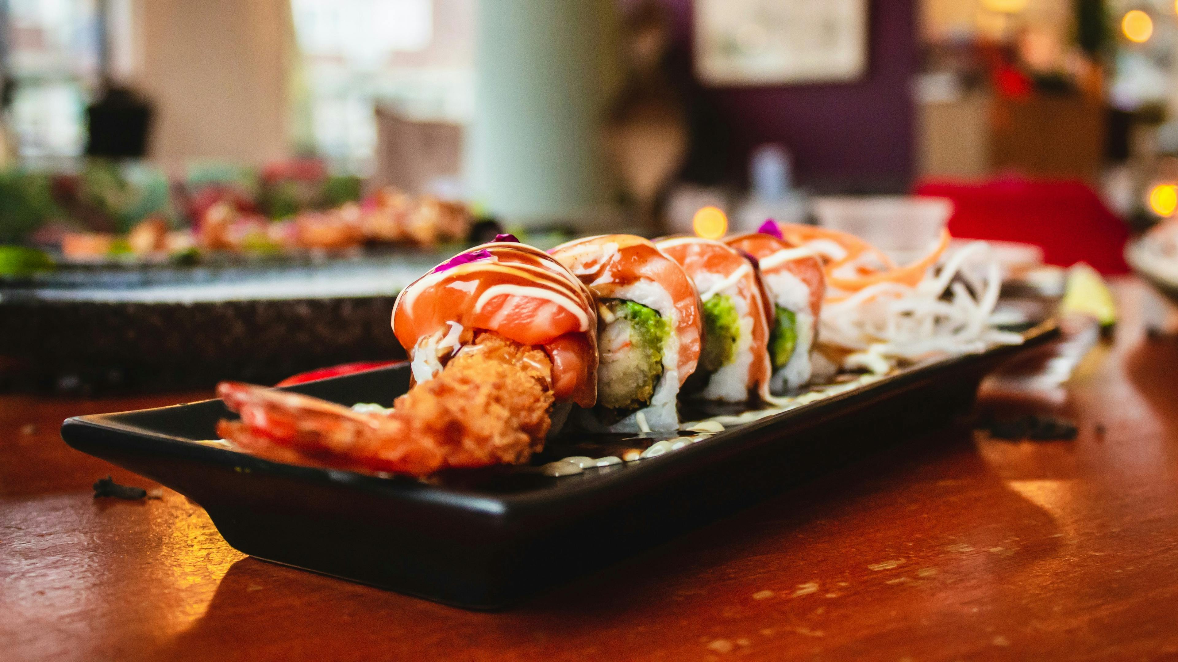 A long plate of sushi rolls with tempura shrimp and sauce is sitting on a wooden table in a restaurant. The background is blurred, showing the warm, inviting ambiance of the space.