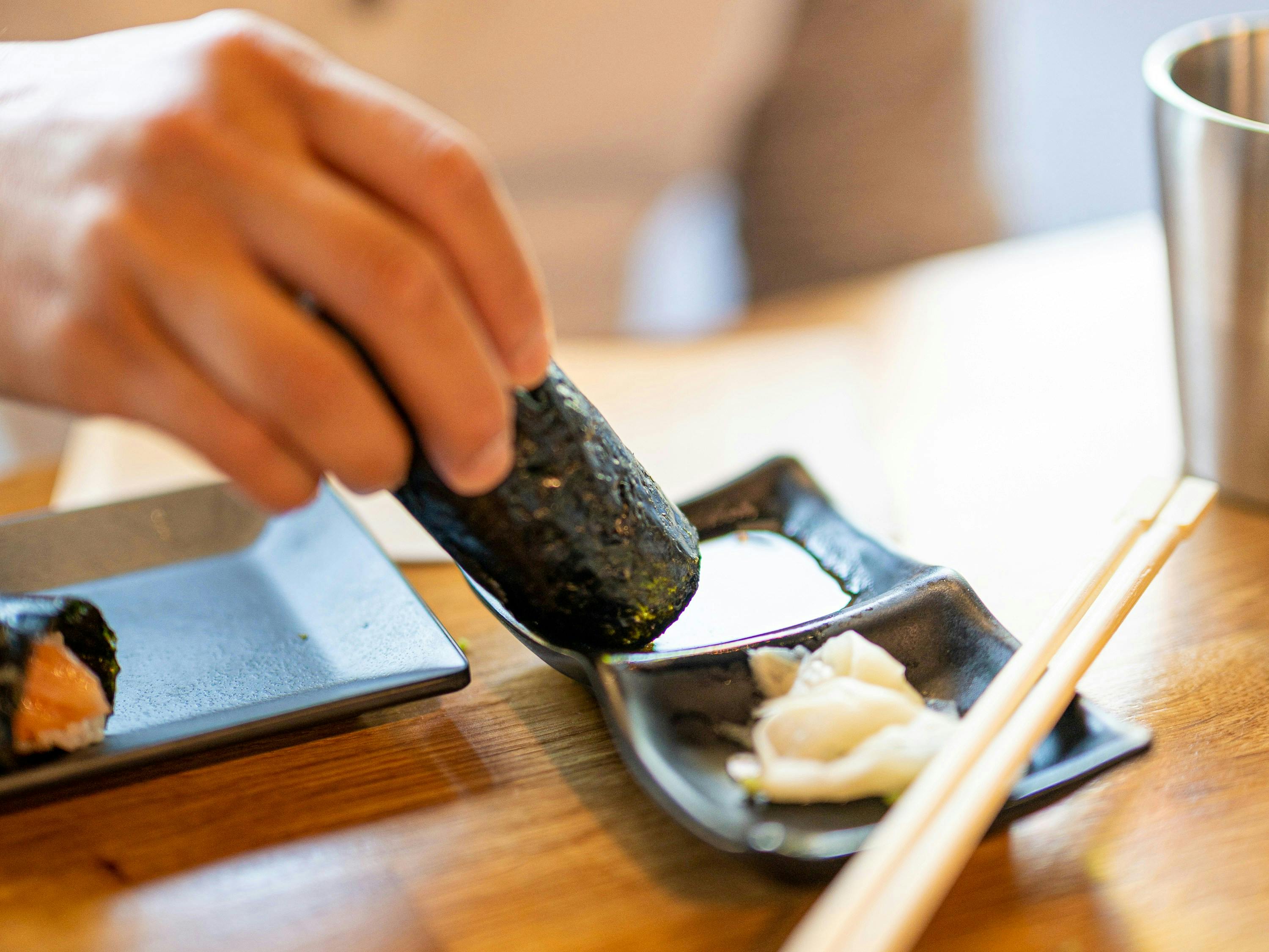 A close-up shot of a person dipping a large sushi roll wrapped in seaweed into a small dish of soy sauce. A small pile of pickled ginger is next to the soy sauce.