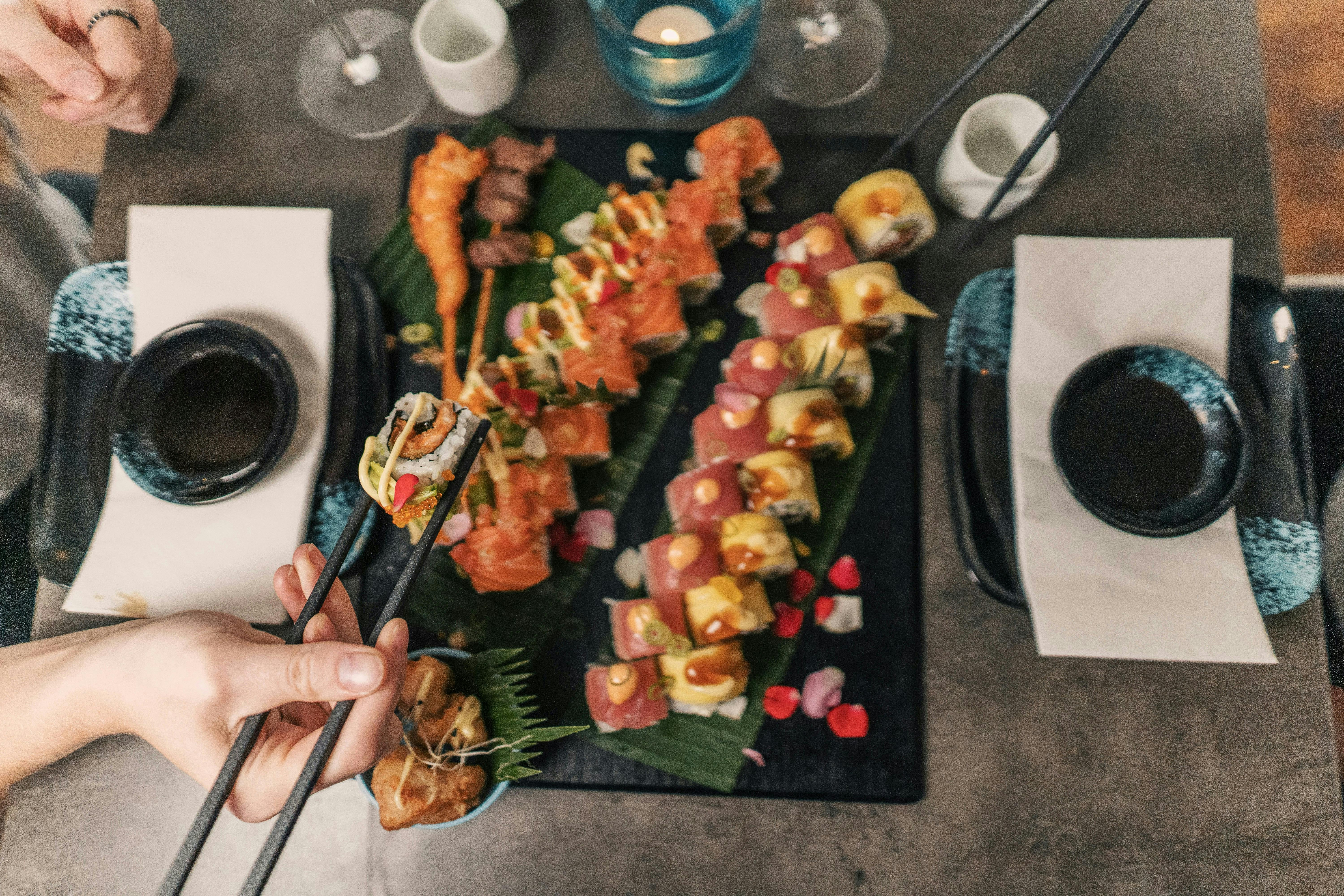 An overhead view of a large platter of assorted sushi rolls, tempura, and other Japanese food on a dark surface. A person is using chopsticks to pick up a piece of sushi.