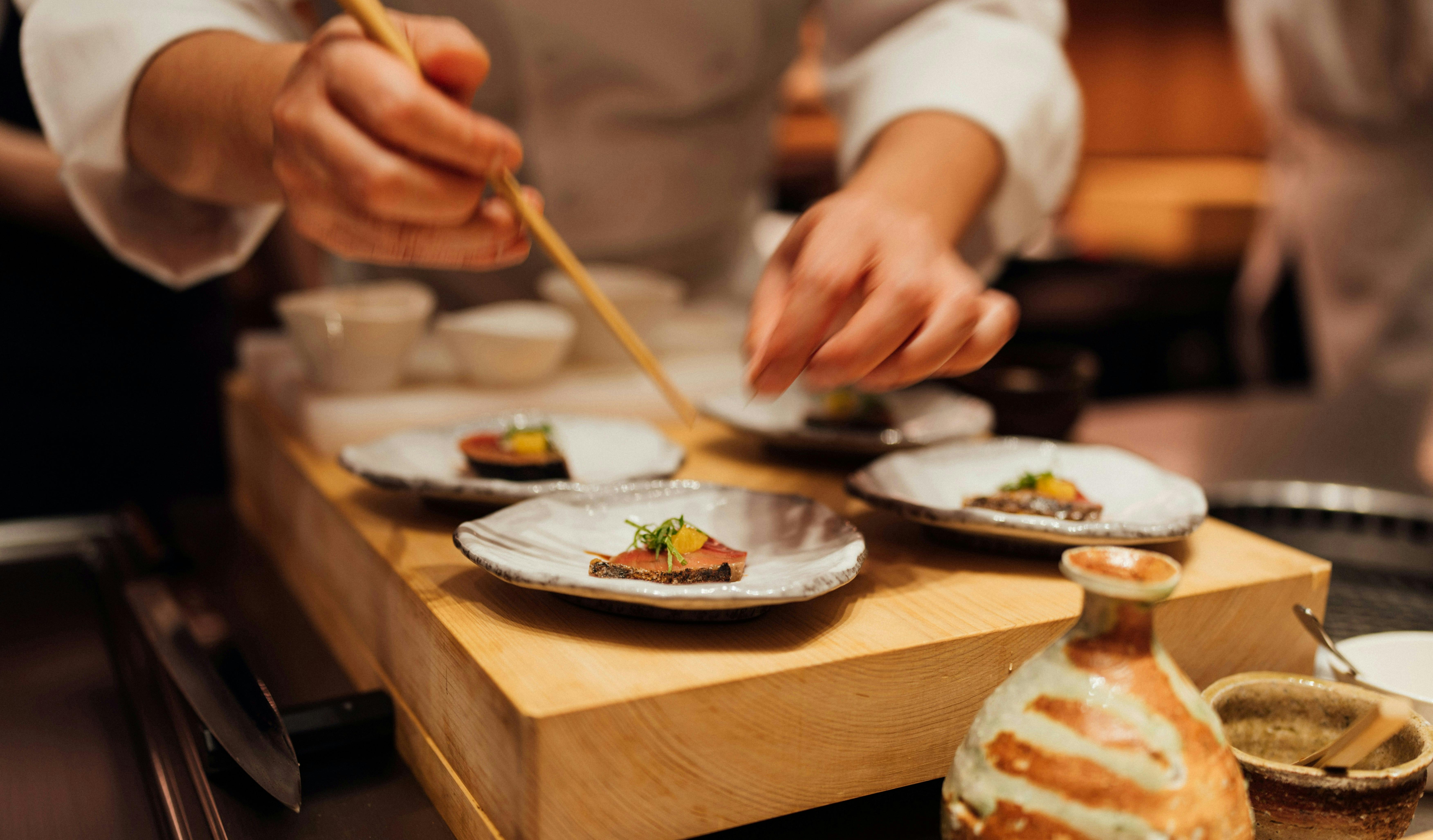 A sushi chef is carefully plating individual pieces of sushi with chopsticks on small, decorative plates on a wooden board. A ceramic sake bottle is in the foreground.
