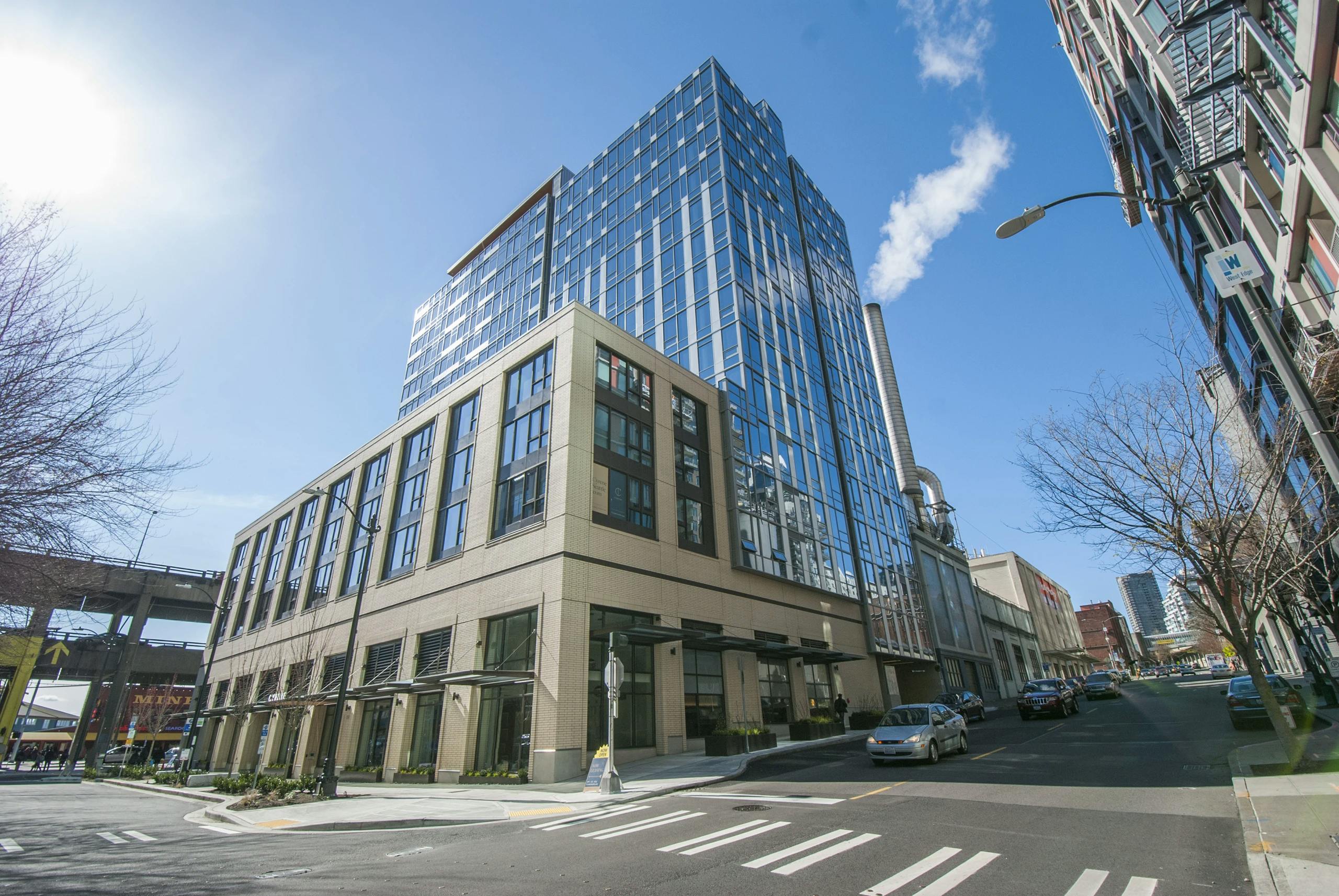 Street-level view of AMLI Waterfront apartment building with glass facade, tan brick, and a clear blue sky above Seattle.