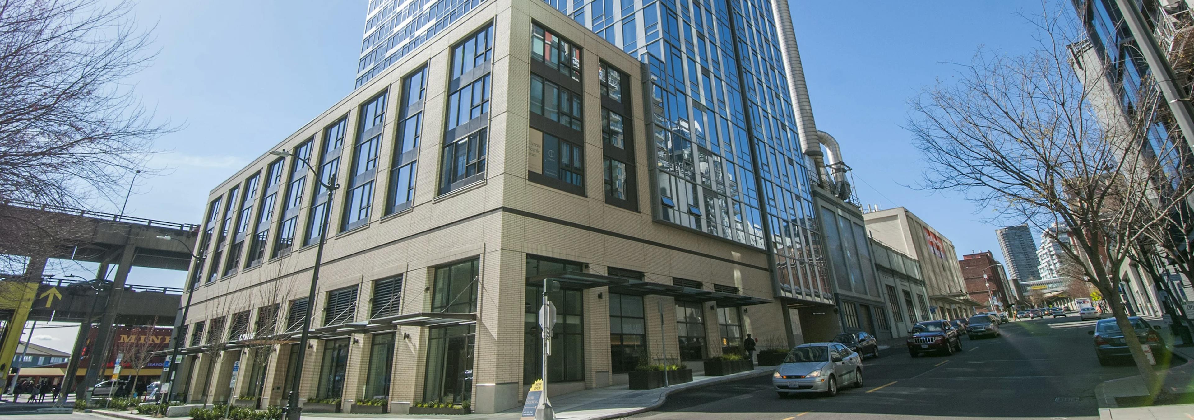 Street-level view of AMLI Waterfront apartment building with glass facade, tan brick, and a clear blue sky above Seattle.