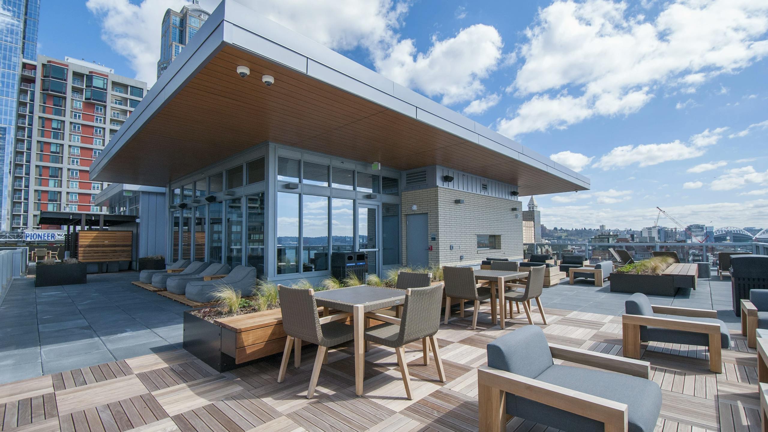 Rooftop terrace at AMLI Waterfront apartments with patio tables, planters, skyline backdrop, and entertainment room.