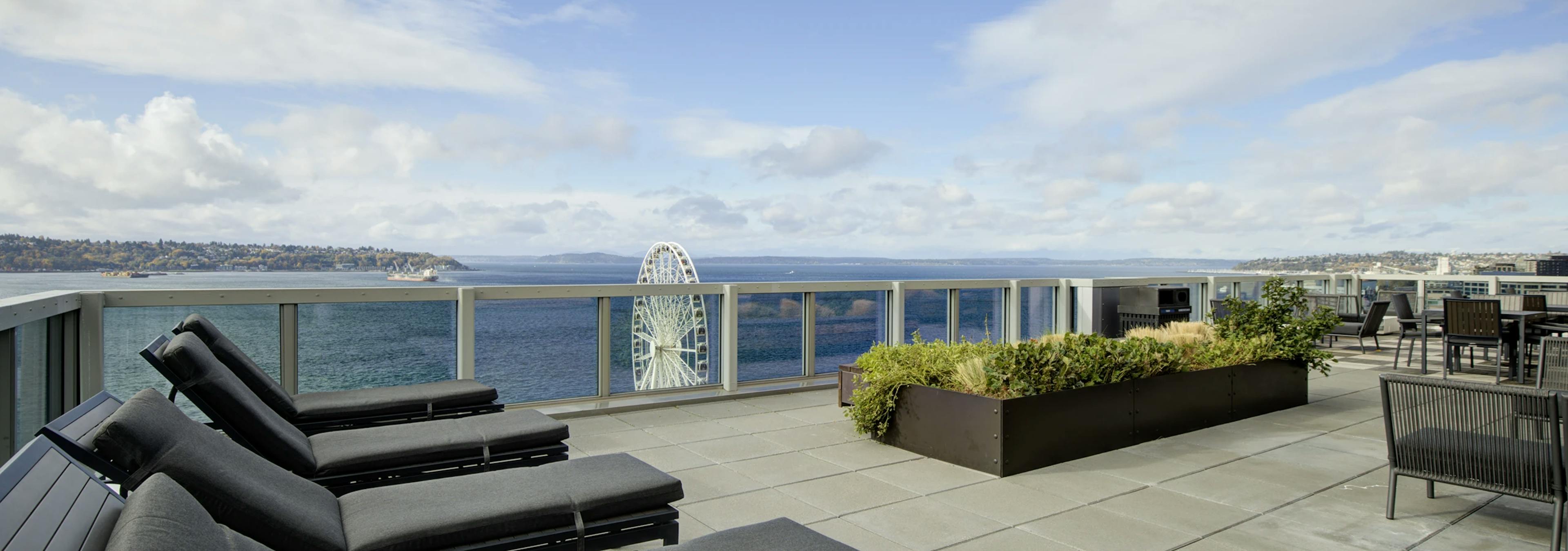 Rooftop deck with gray loungers, planter beds, and Puget Sound views on a sunny day at AMLI Waterfront apartments