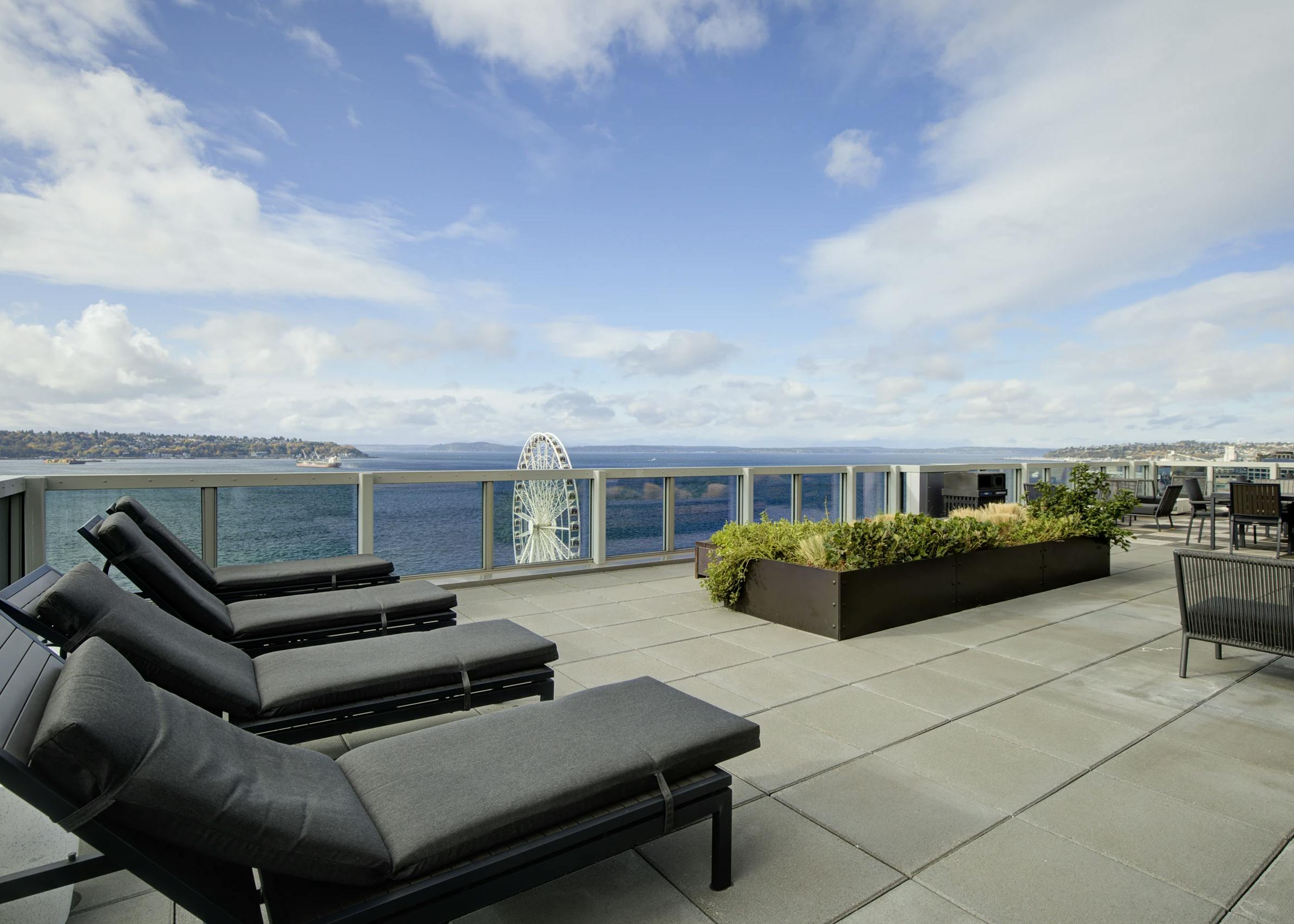 Rooftop deck with gray loungers, planter beds, and Puget Sound views on a sunny day at AMLI Waterfront apartments