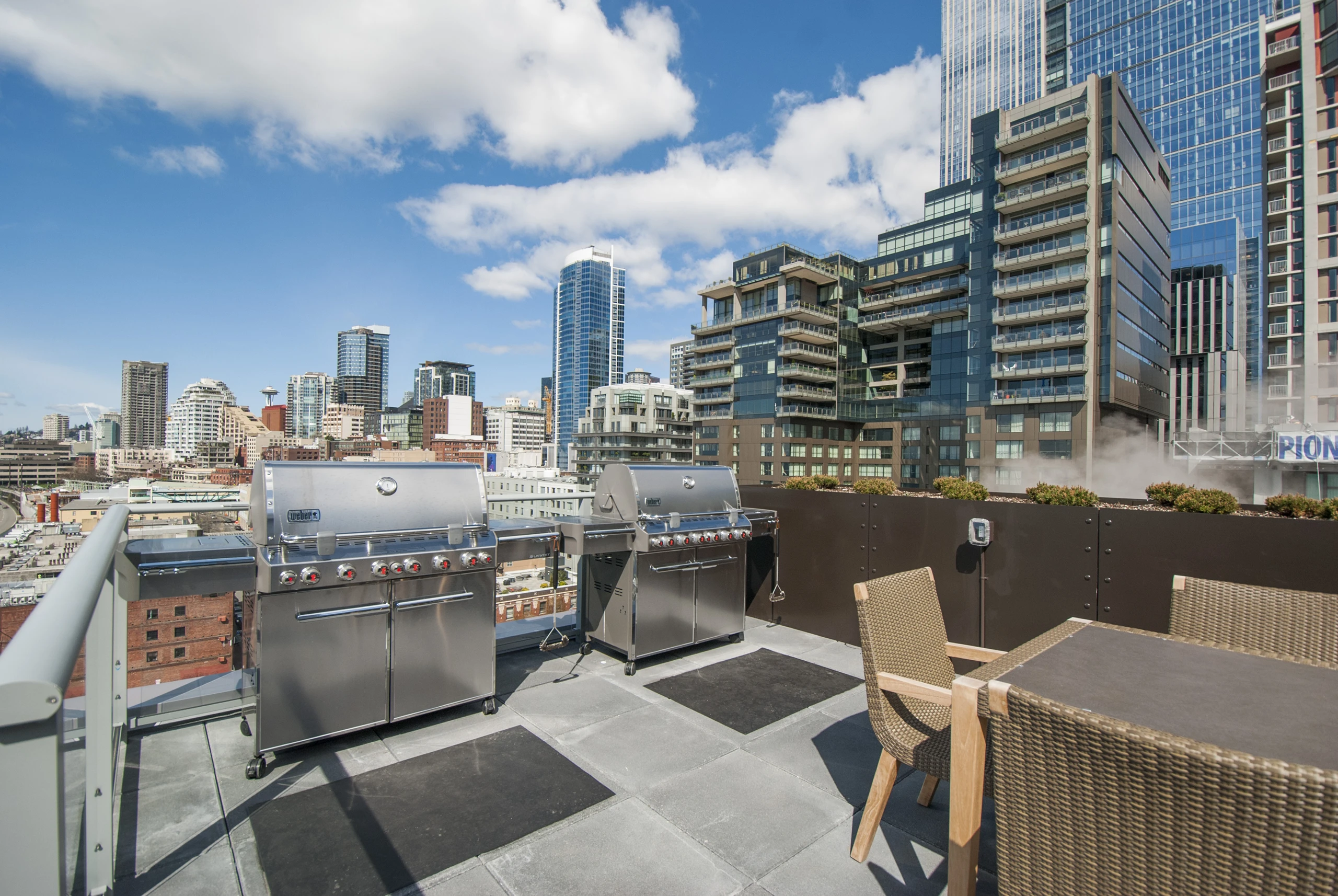 Rooftop grill area at AMLI Waterfront apartments with stainless steel grills, woven patio chairs, and downtown skyline views.