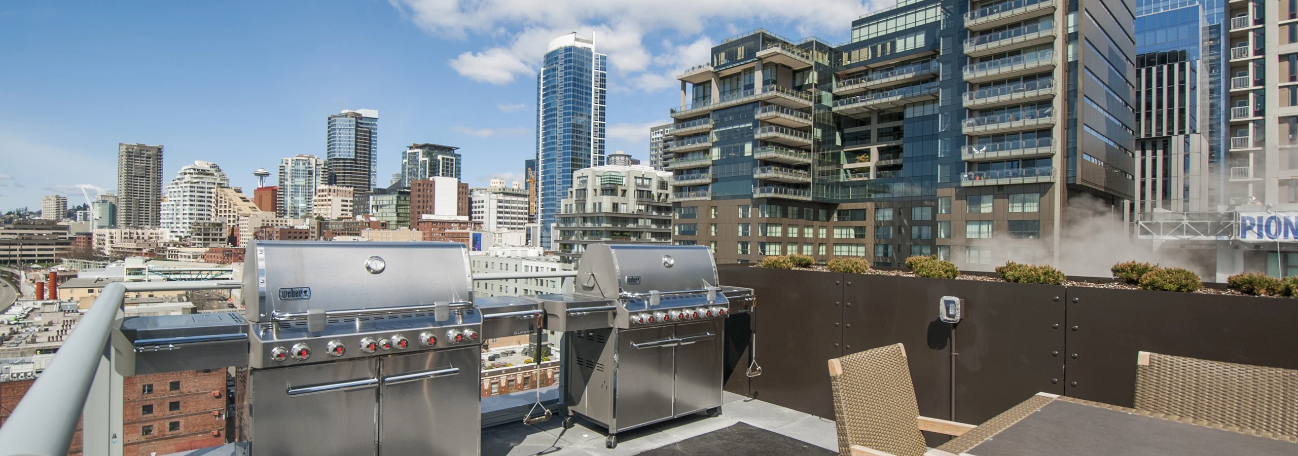 Rooftop grill area at AMLI Waterfront apartments with stainless steel grills, woven patio chairs, and downtown skyline views.
