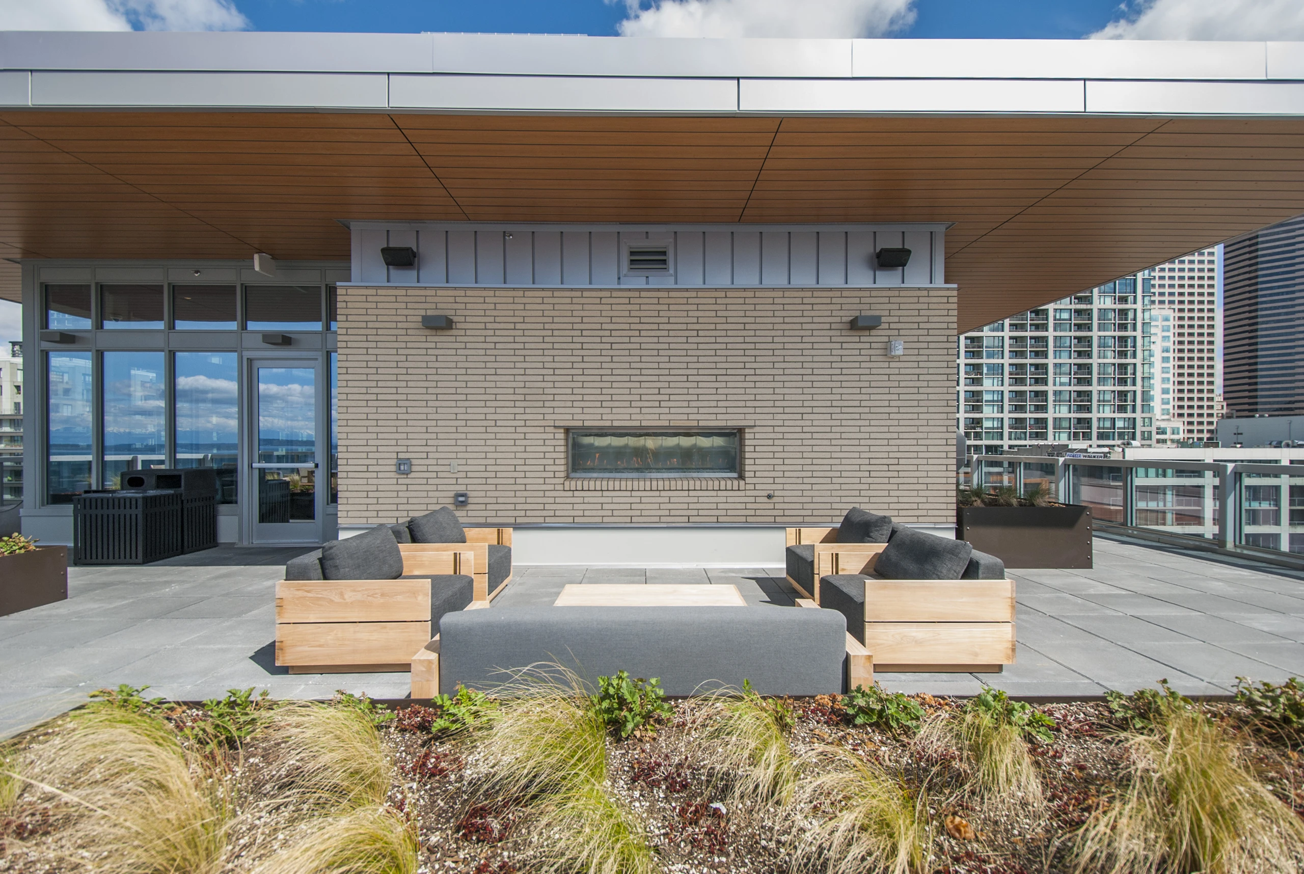 Outdoor fireplace lounge at AMLI Waterfront apartments with cushioned wood seating, beige brick facade, and glass railings.