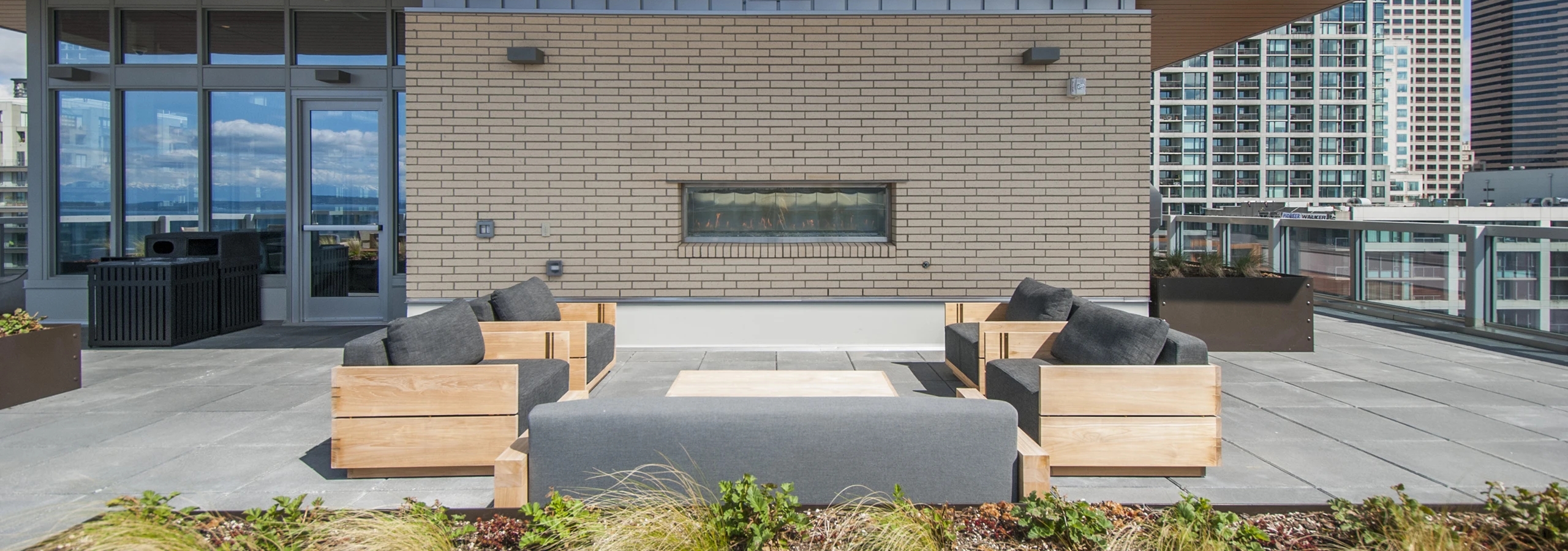 Outdoor fireplace lounge at AMLI Waterfront apartments with cushioned wood seating, beige brick facade, and glass railings.