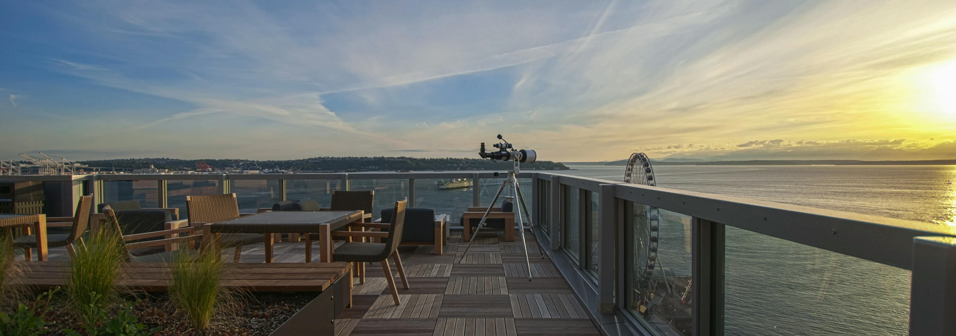 AMLI Waterfront rooftop deck with wood furniture, telescope, and sunset views over the bay and Seattle Great Wheel.