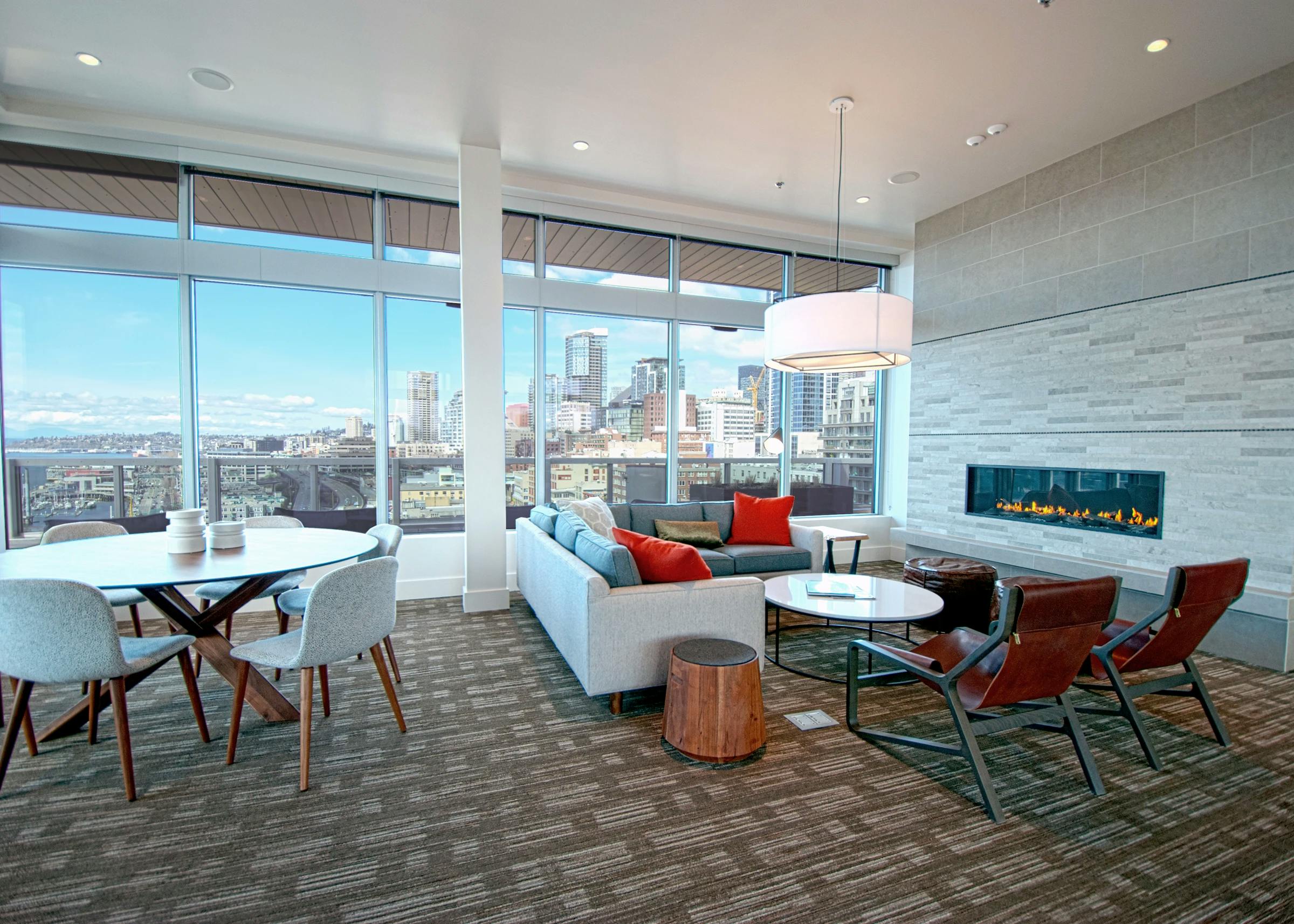 Rooftop clubroom at AMLI Waterfront apartments with sectional sofa, fireplace, leather chairs, and skyline views through tall windows.