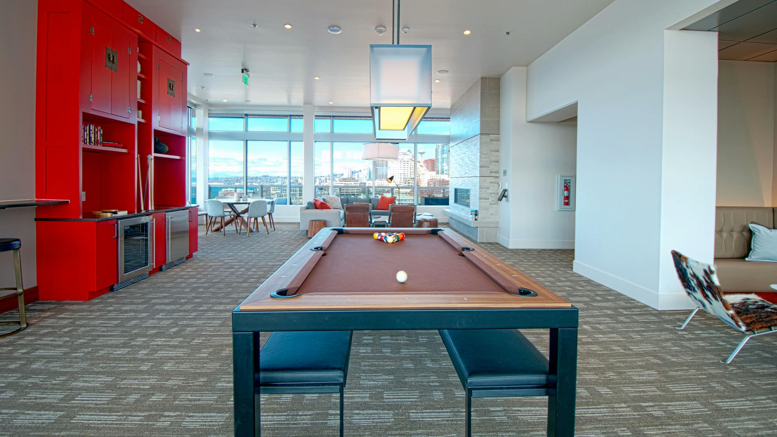 Clubroom game area at AMLI Waterfront apartments with red cabinetry, pool table, lounge seating, and large windows overlooking the city.