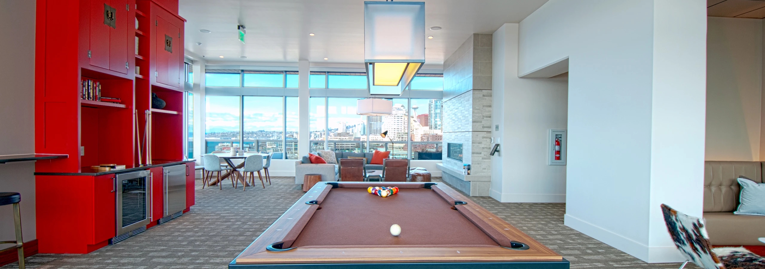 Clubroom game area at AMLI Waterfront apartments with red cabinetry, pool table, lounge seating, and large windows overlooking the city.
