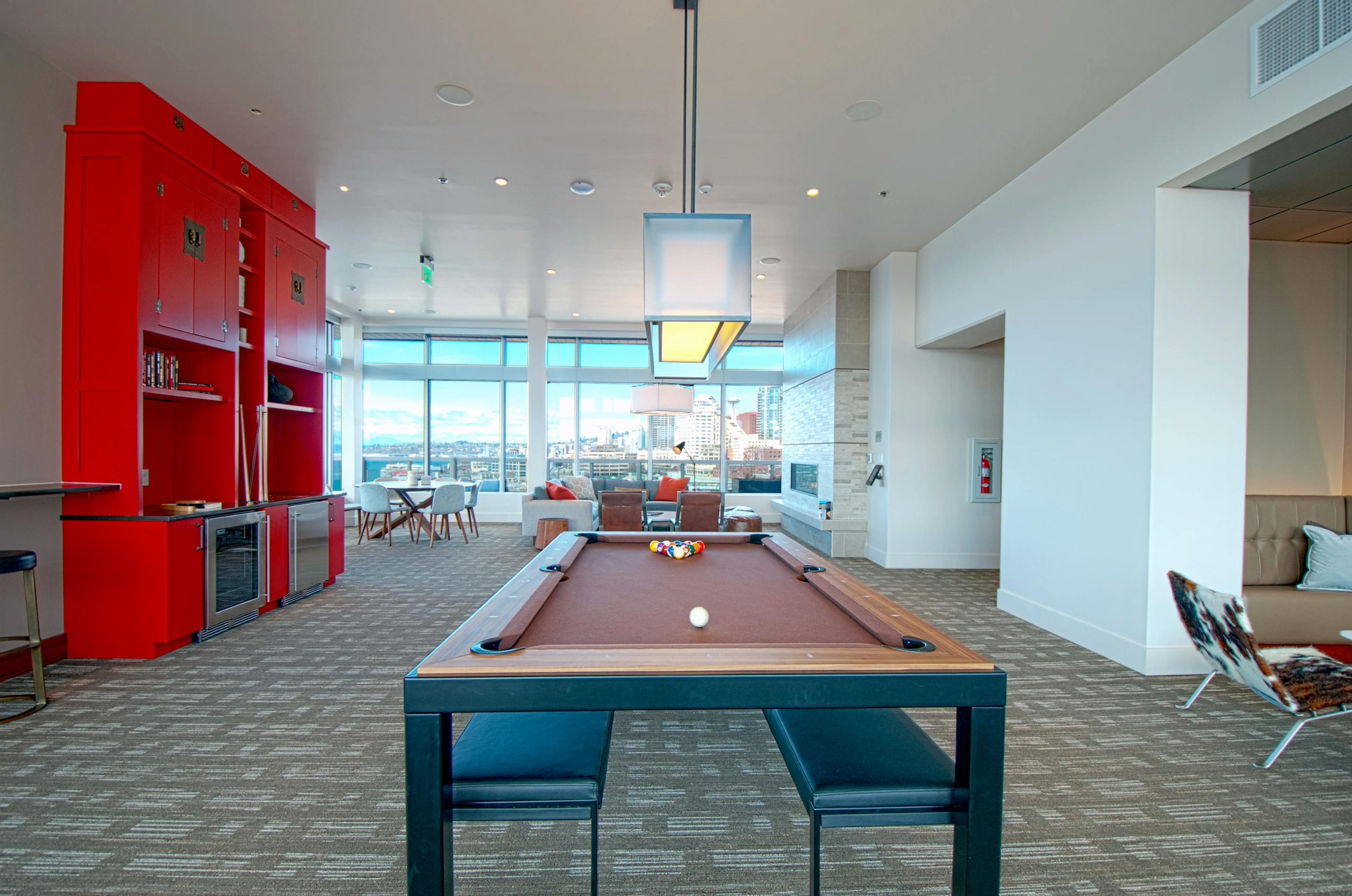 Clubroom game area at AMLI Waterfront apartments with red cabinetry, pool table, lounge seating, and large windows overlooking the city.