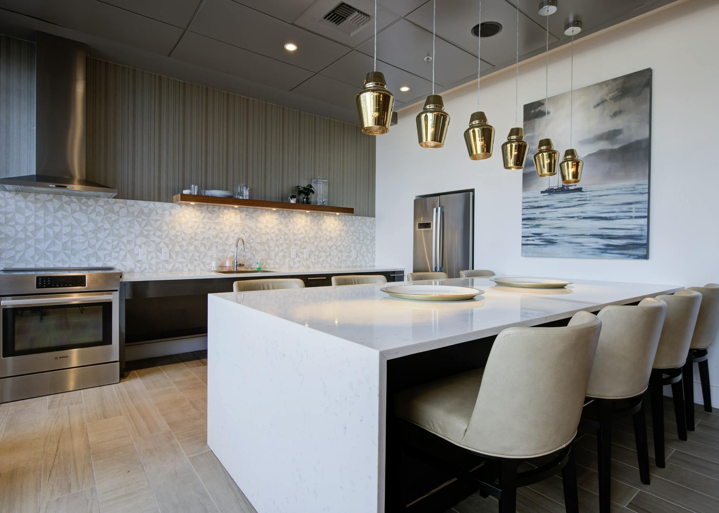 AMLI Waterfront clubroom kitchen at AMLI with quartz island, bar stools, gold pendant lighting, and full backsplash.